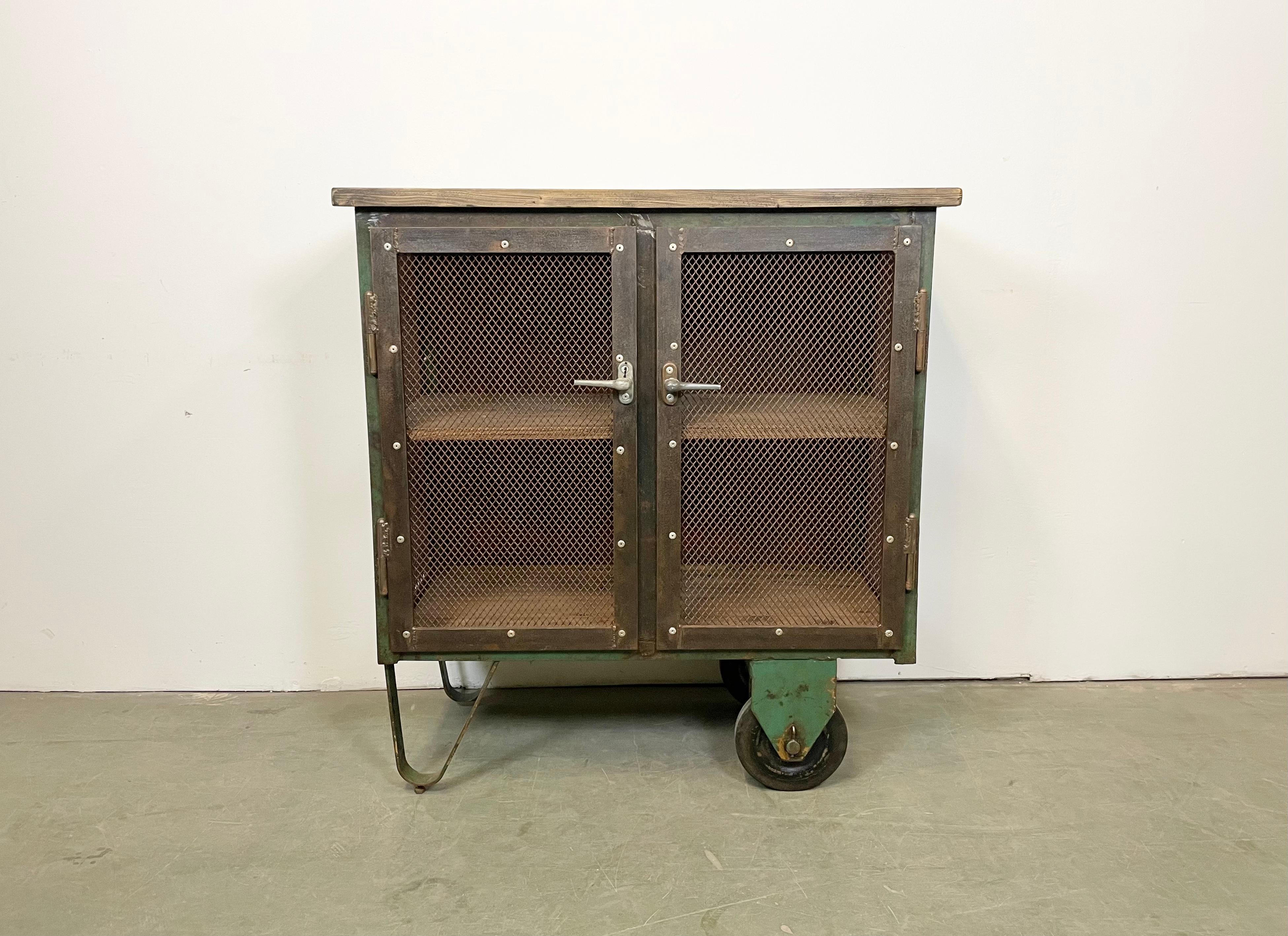 Iron cabinet from the 1960s it features a green iron construction with mesh doors, two wooden shelwes, a wooden top and two original wheels. The weight of the cabinet is 73 kg.