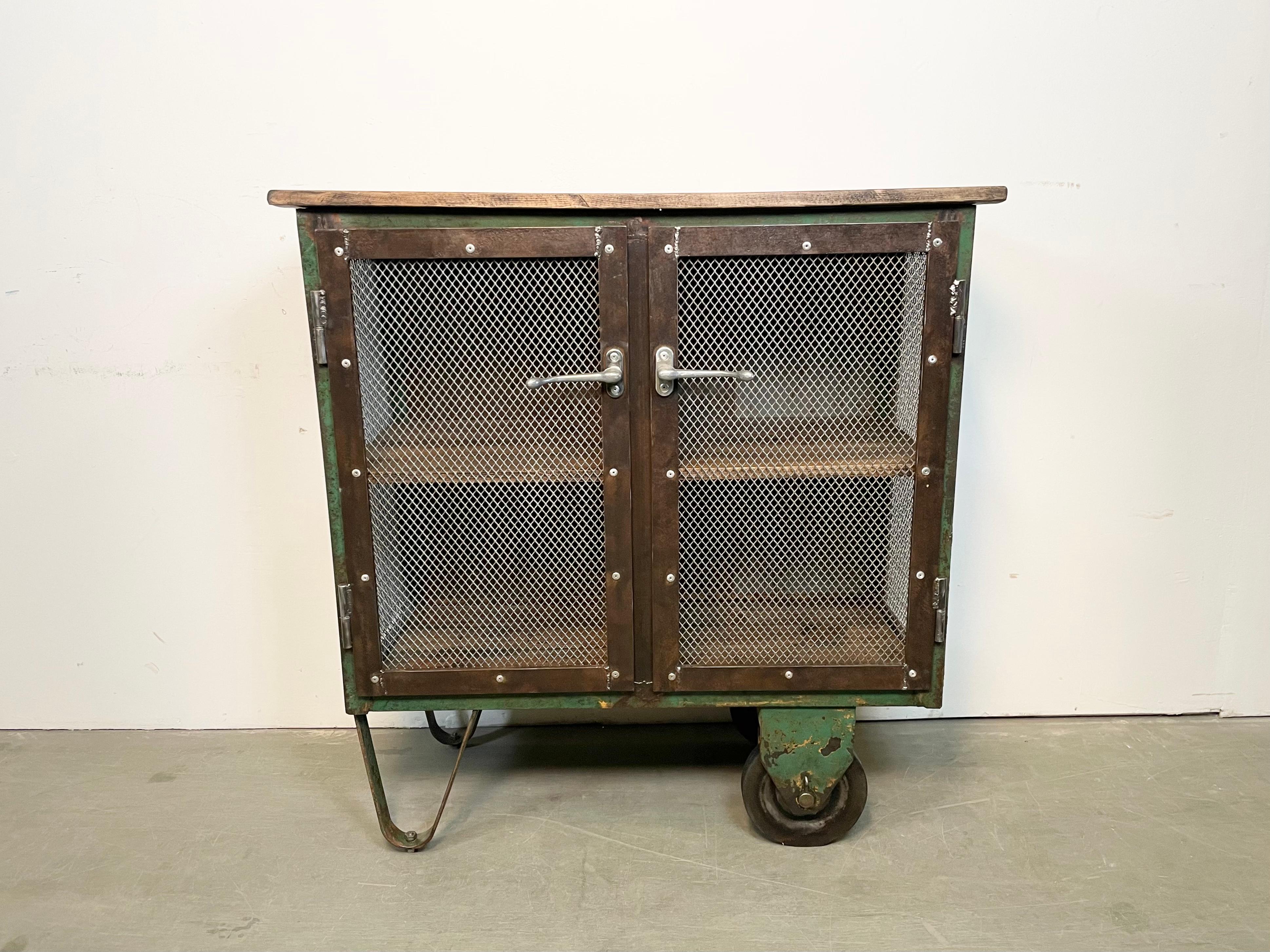 Iron cabinet from the 1960s it features a green iron construction with mesh doors, two wooden shelwes, a wooden top and two original wheels. The weight of the cabinet is 65 kg.