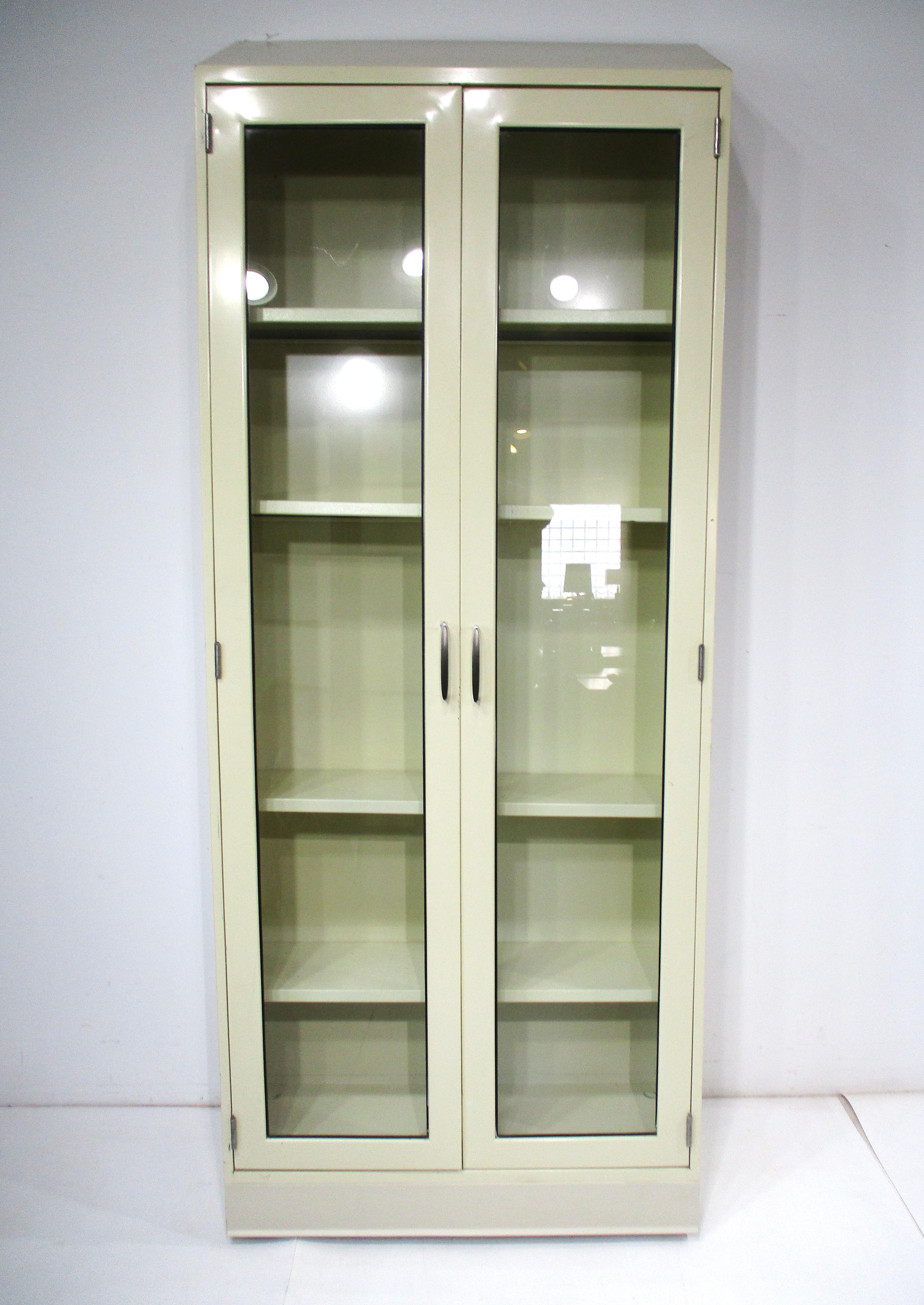 A nice streamline industrial metal bookcase / cabinet in a medium cream color with four adjustable matching shelves having two glass doors with stainless steel handles . The taller height of the cabinet peaks to the back of the top giving it a nice