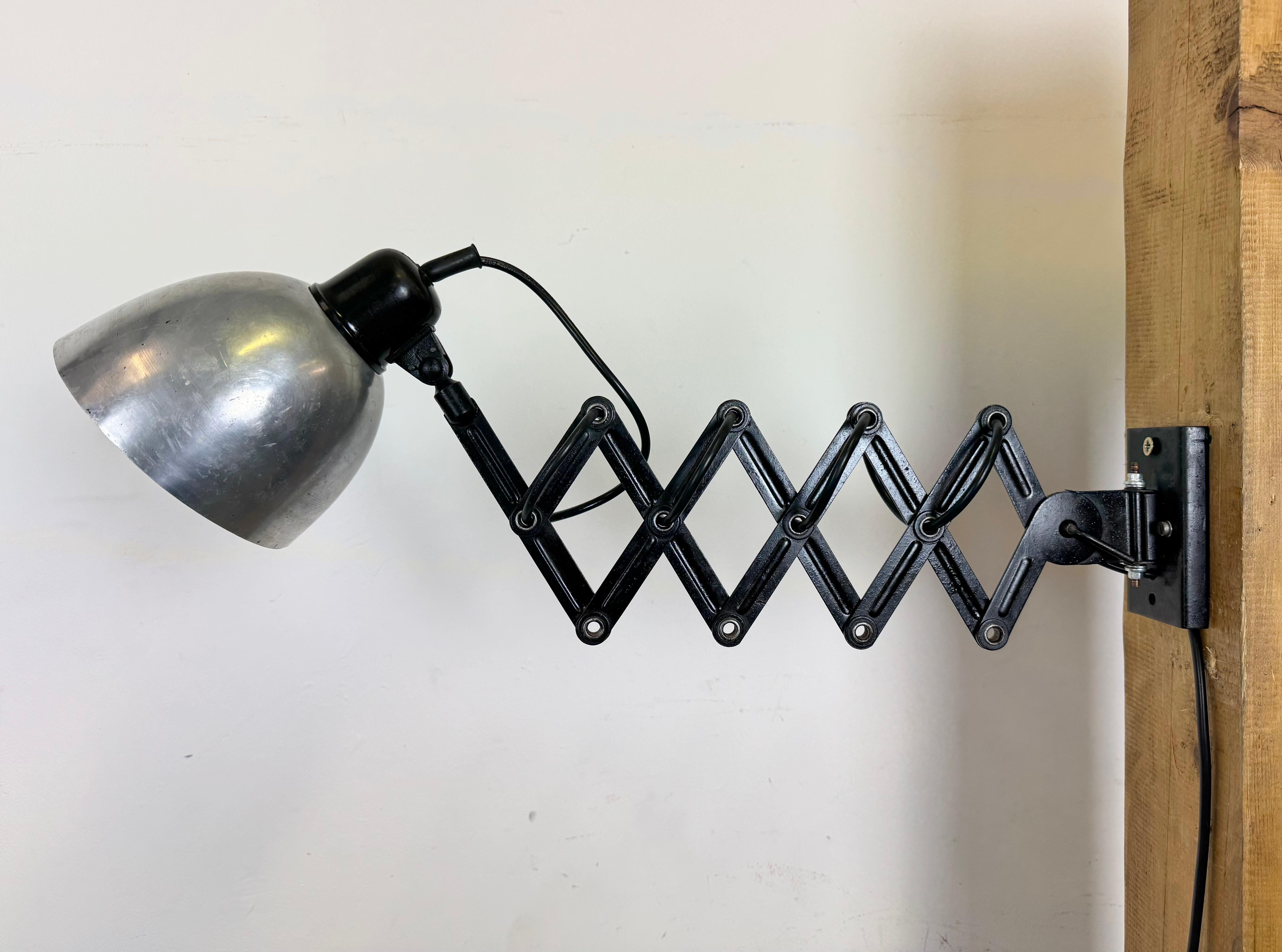 Cette applique industrielle vintage à ciseaux a été produite dans l'ancienne Tchécoslovaquie au cours des années 1950. La lampe est dotée d'un abat-jour en métal argenté et d'un couvercle en bakélite. Le bras de ciseaux en fer noir est extensible et