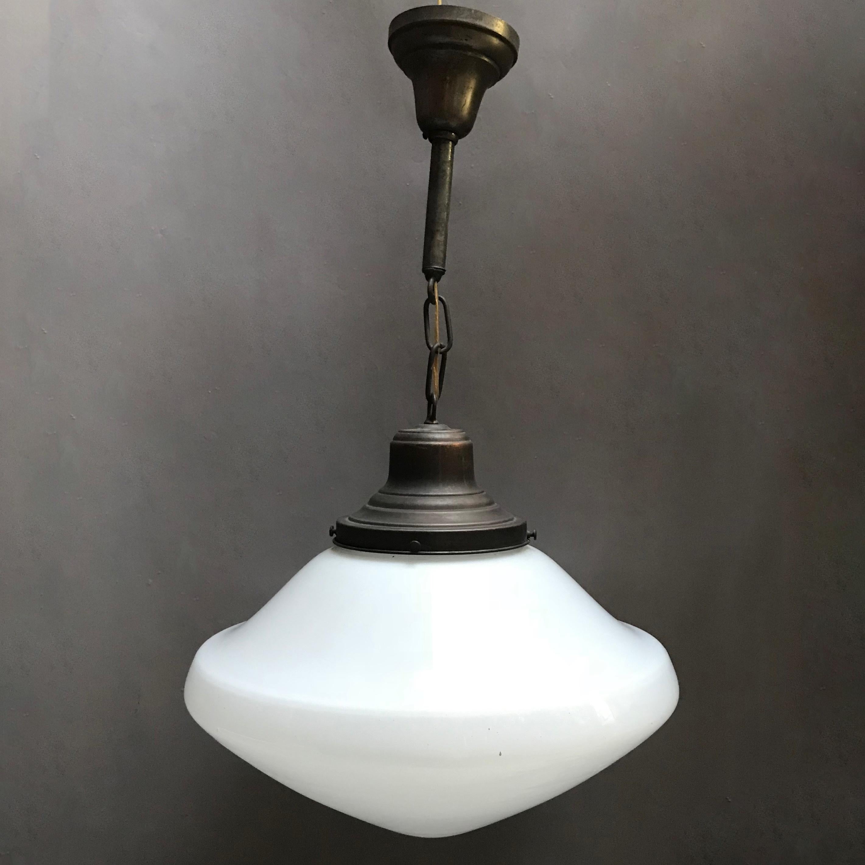 Early 20th century, industrial, pharmacy, pendant light features a peaked and contoured milk glass shade with patinated brass fitter, chain and canopy, is wired to accept up to a 200 watt bulb. The total height of the pendant is 24 inches, the