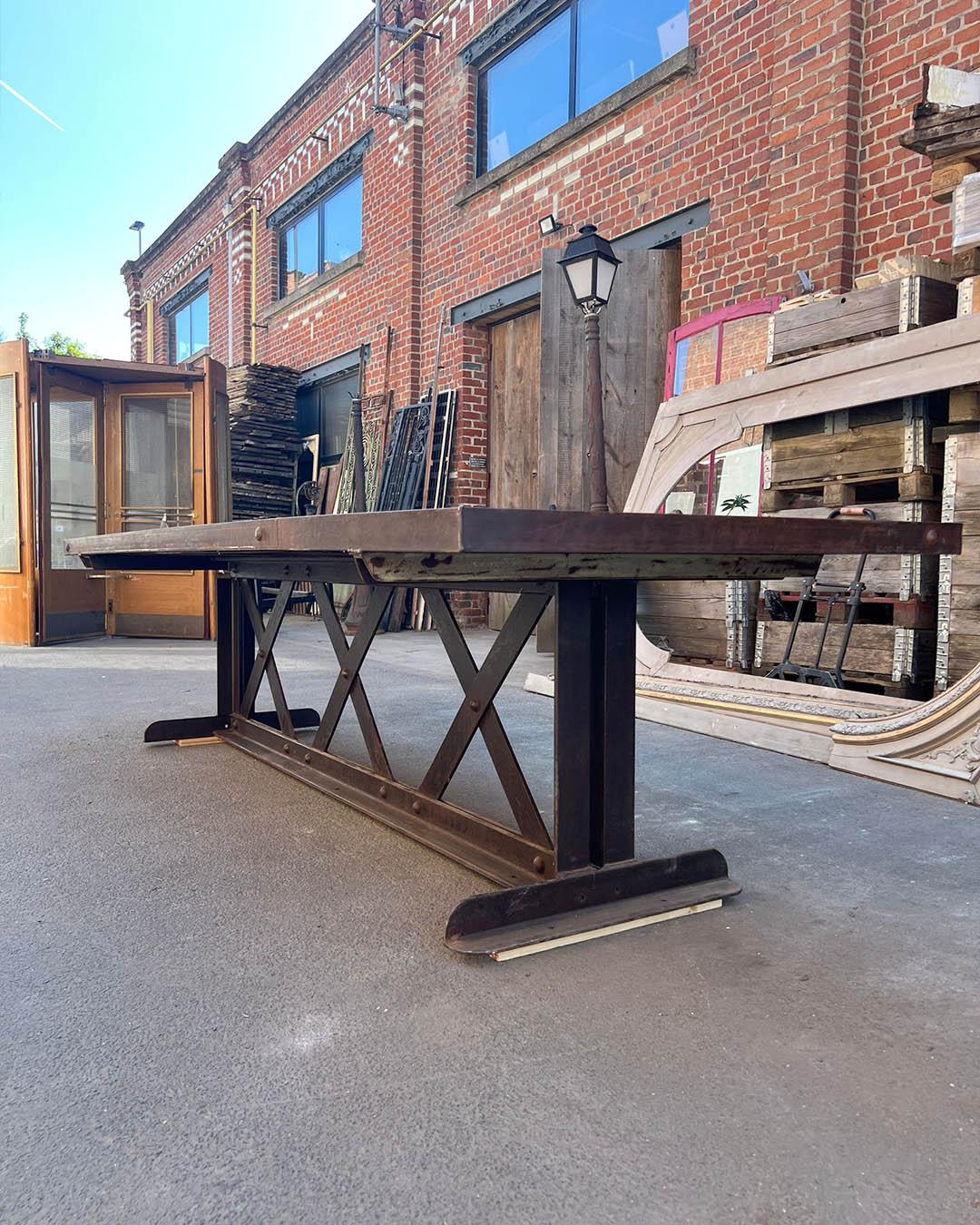 Imponente mesa de estilo industrial con un grueso tablero de roble macizo que descansa sobre una robusta base de hierro fundido con detalles remachados en forma de X. La armoniosa combinación de madera en bruto y metal envejecido confiere a esta
