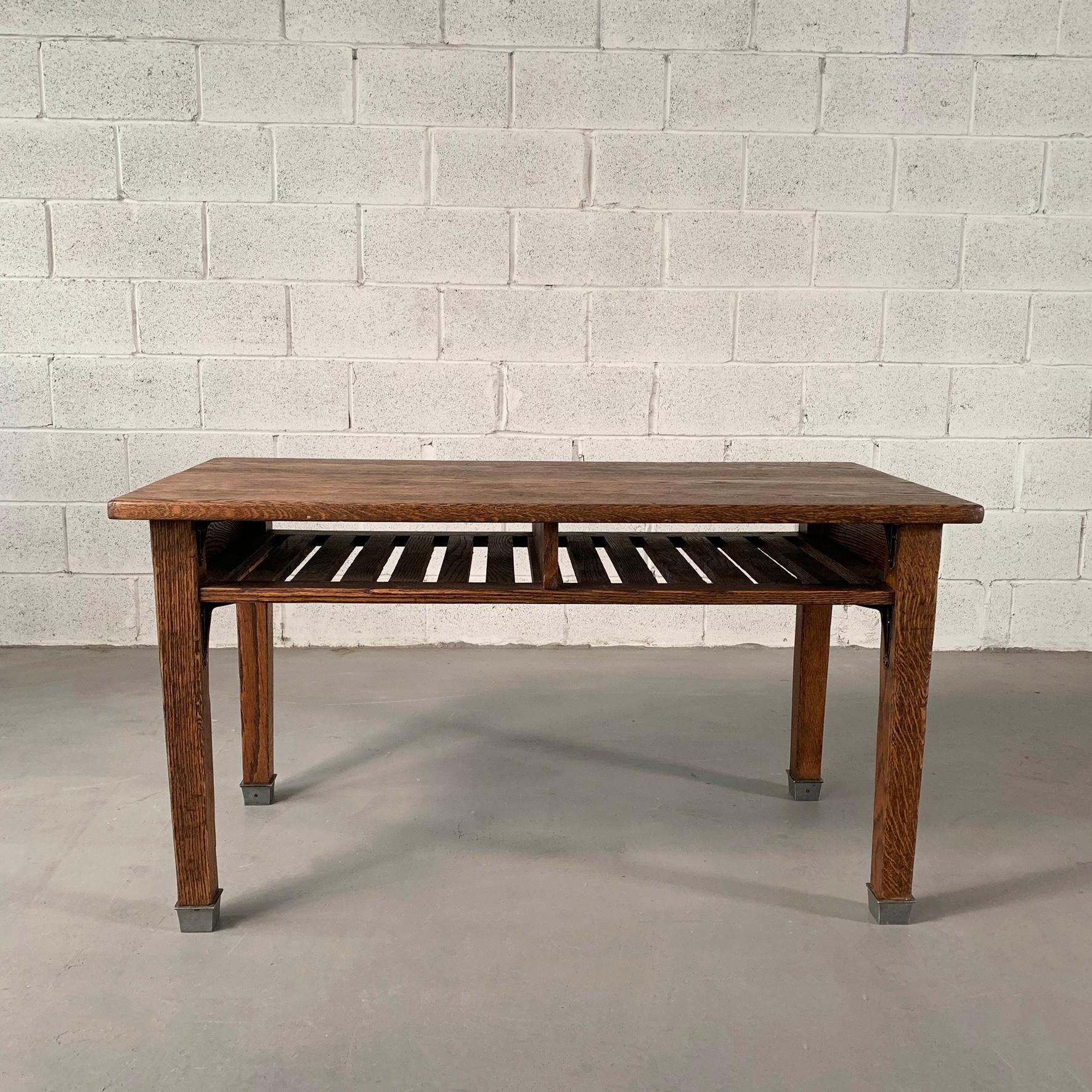 Industrial, 1940s, quarter sawn oak, school, library table or desk features a 4 inch high, slat-bottom, under shelf for books open on both sides with steel capped legs. 2 tables are available. The leg height clearance is 19.5 inches