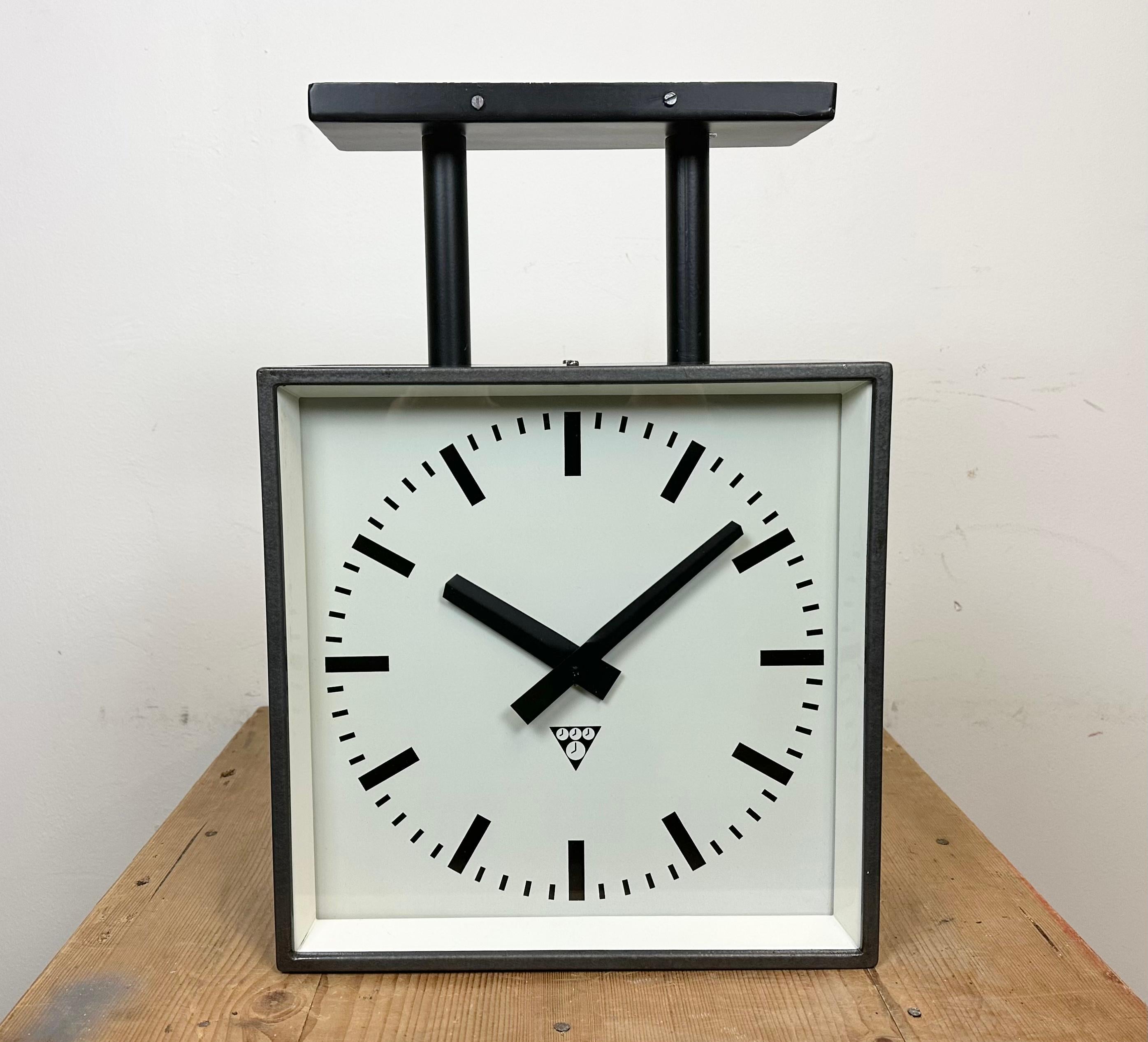 This square double-sided railway, school or factory ceiling clock was produced by Pragotron, in former Czechoslovakia, during the 1970s. The piece features a black metal body, a dark grey hammerpaint clock frames, a clear glass covers and an