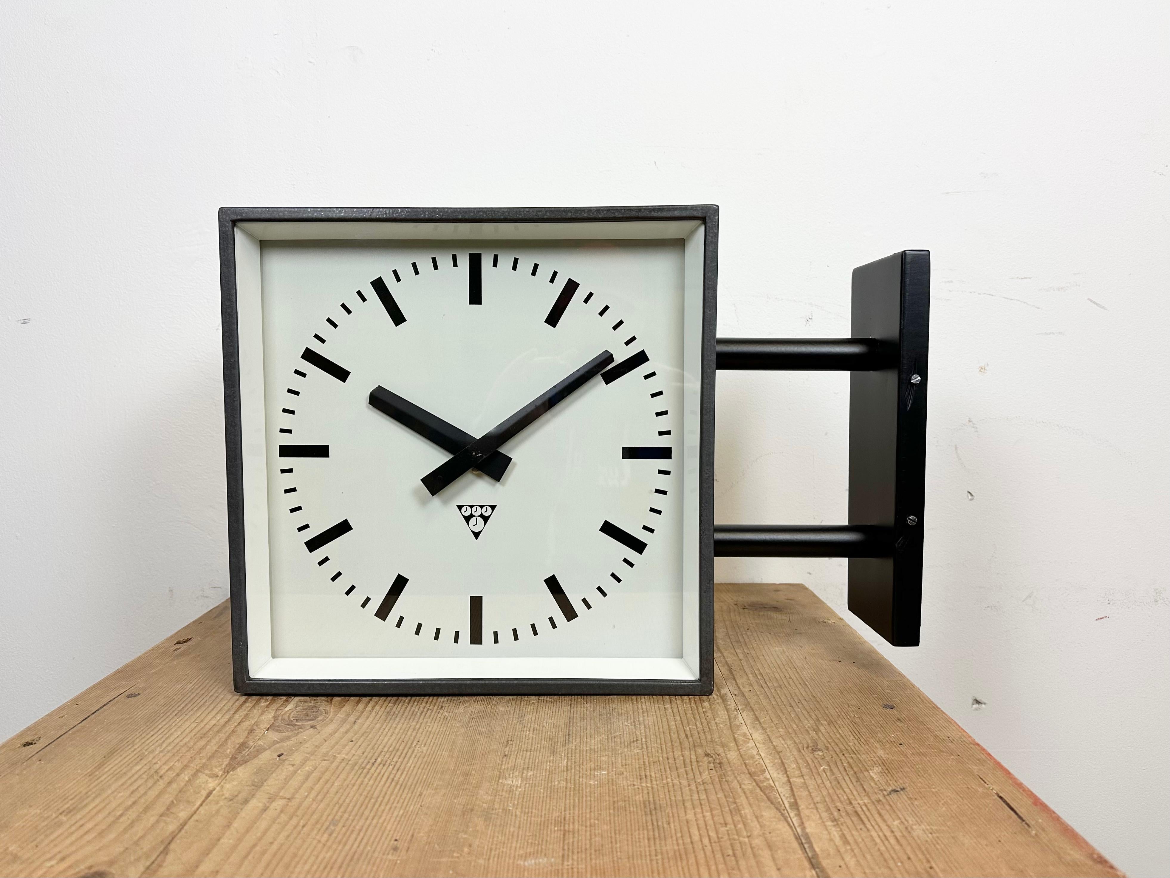 This square double-sided railway, school or factory wall clock was produced by Pragotron, in former Czechoslovakia, during the 1970s. The piece features a black metal body, a dark grey hammerpaint clock frames, a clear glass cover, and an aluminum