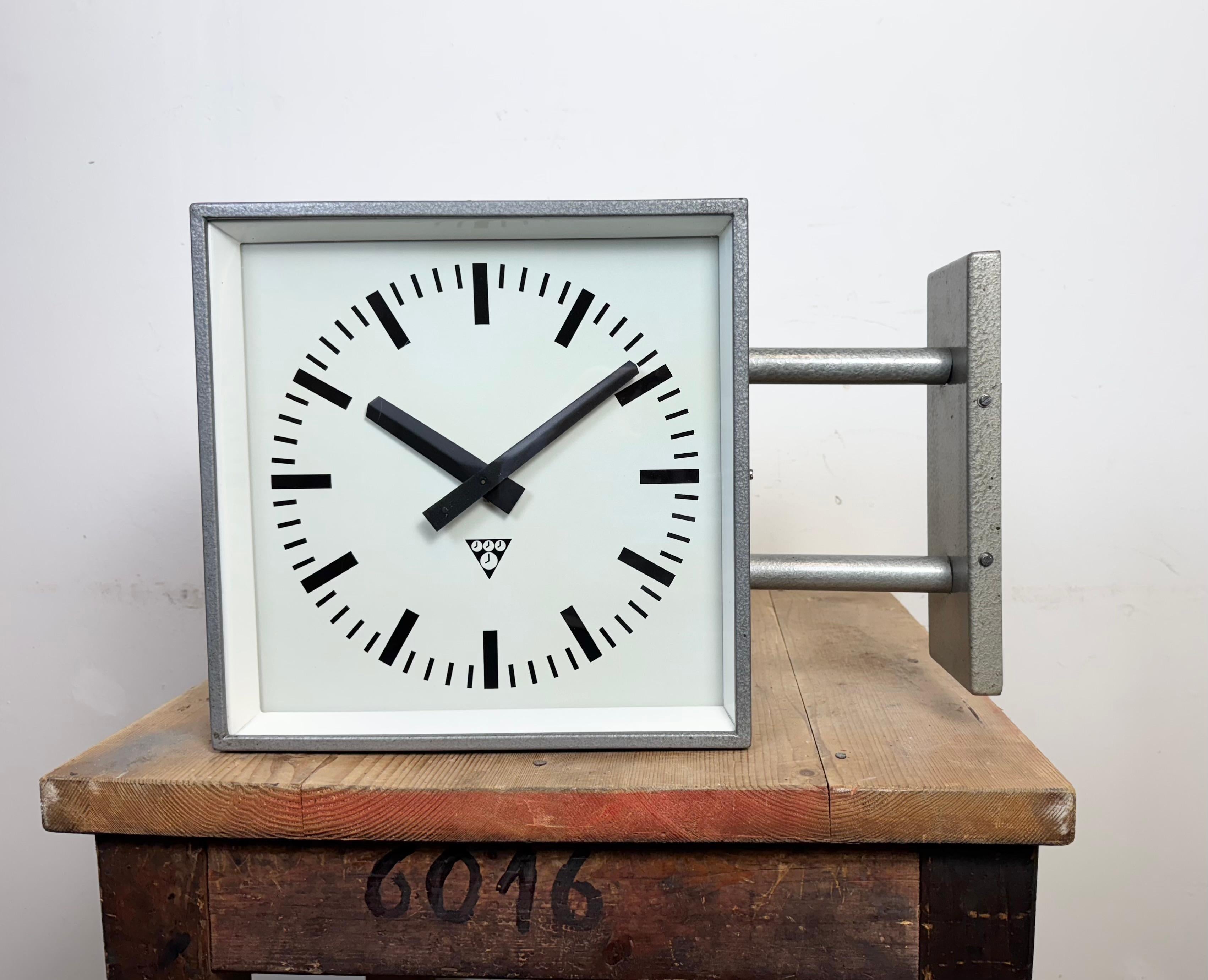 This square double sided railway, school or factory wall clock was produced by Pragotron, in former Czechoslovakia, during the 1970s. The piece features a grey metal body, a grey hammer paint clock frames, a clear glass covers and an aluminum dials
