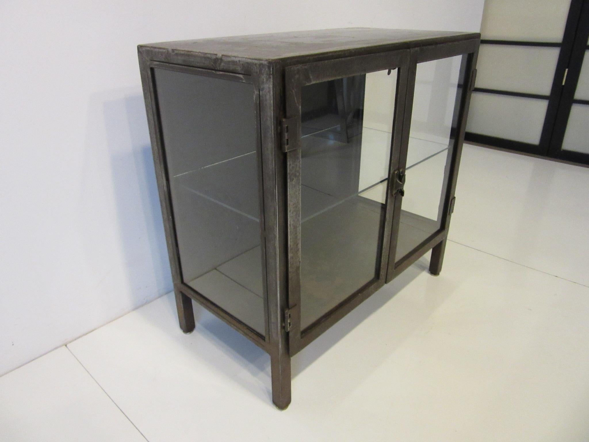A smaller sized Industrial styled steel display cabinet / bookcase with glass sides and front having two doors a turn latch and one non adjustable shelve. The striped and patina surfaces give the piece a raw and warm look ready for your bathroom,