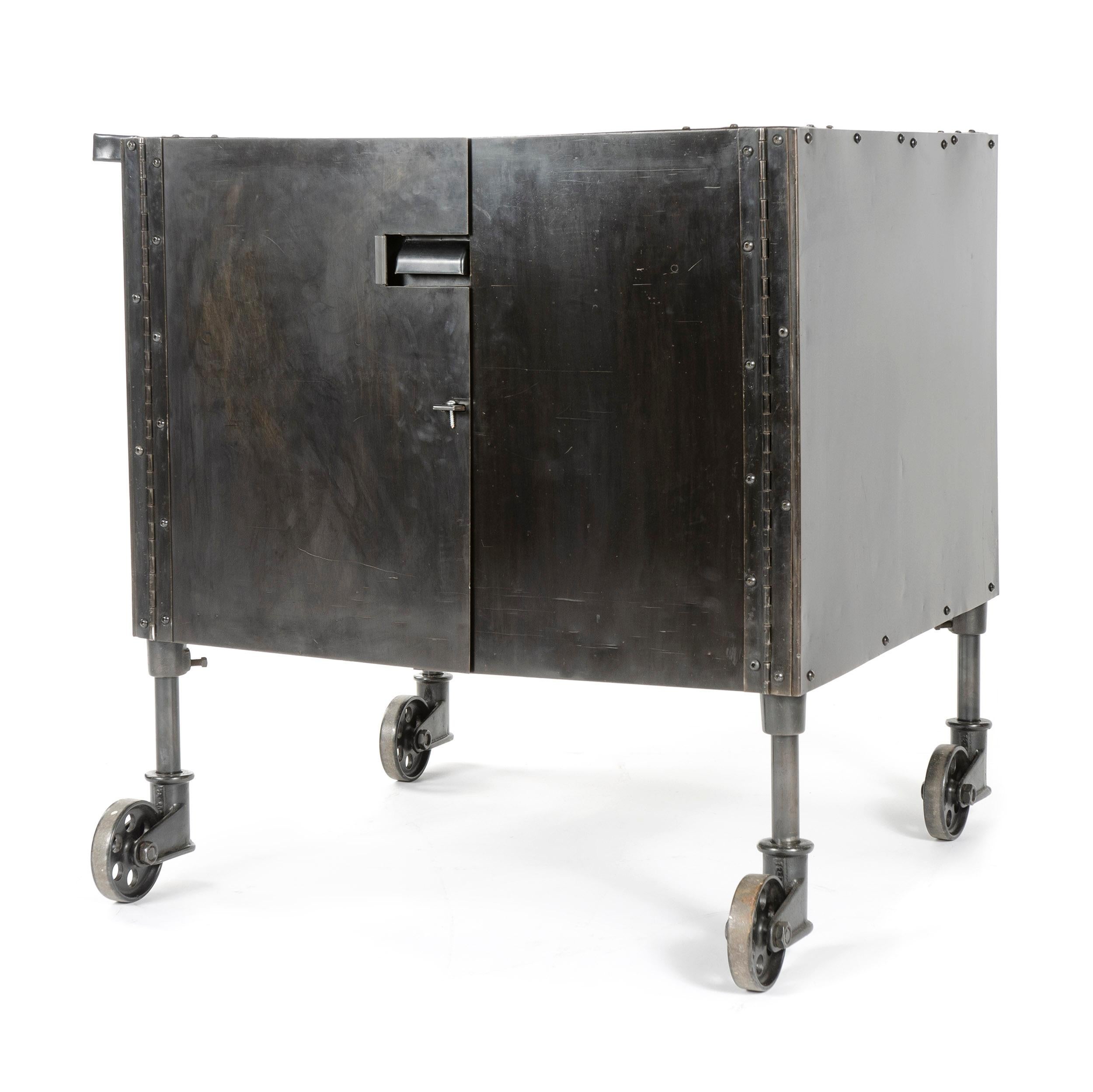A patinated steel industrial cabinet on heavy-duty perforated casters featuring a recessed top, lateral grab bar and two doors opening to a central drawer over open storage. Made in USA, circa 1930s.