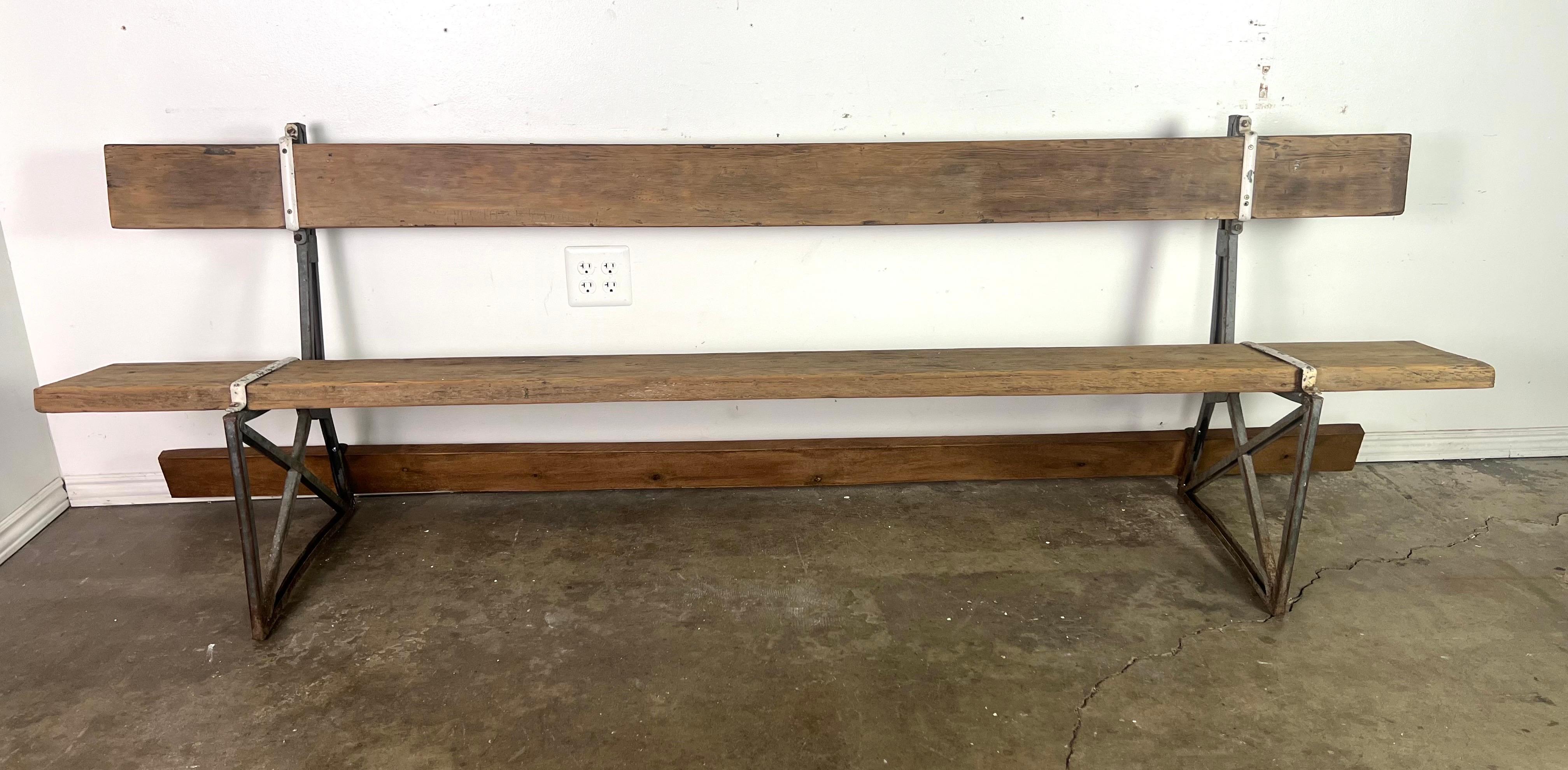 Se trata de un banco rústico de madera con un estilo minimalista e industrial.  El banco tiene un largo asiento y respaldo de madera, ambos con un acabado natural desgastado, que le da un aspecto vintage.  El armazón está formado por soportes