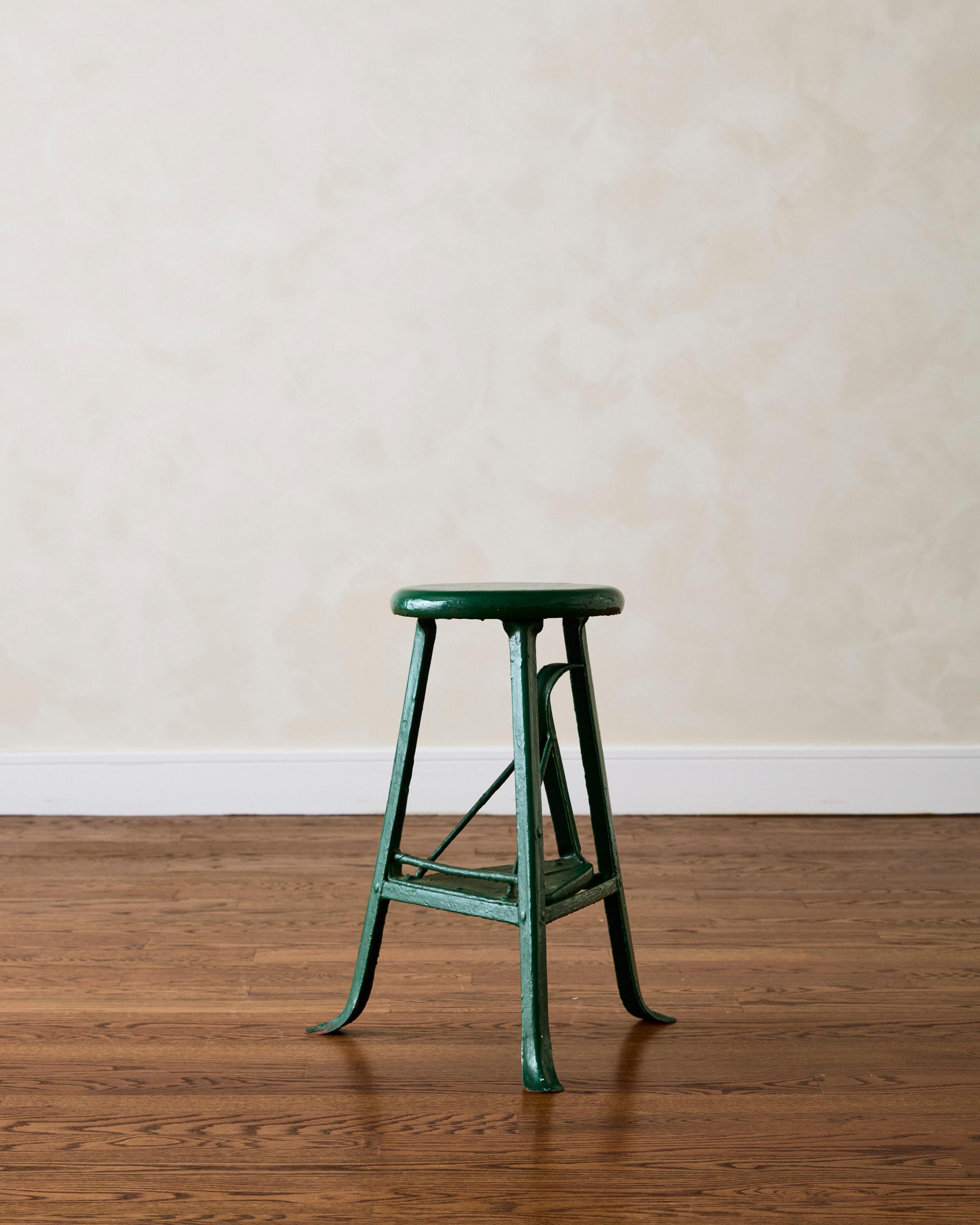 Industrial three-legged stool made of green-painted steel, featuring a swing-out step.

