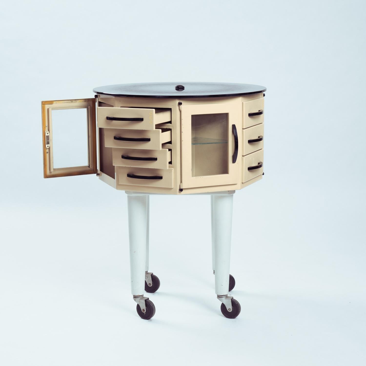 Real dentist’s furniture by Leon Martin, round and rotating, on metal casters; composed of a box articulated on a base around a central axis with a black glass and also turnable top. 
4 doors and 4 series of drawers.
ideal to showcase your