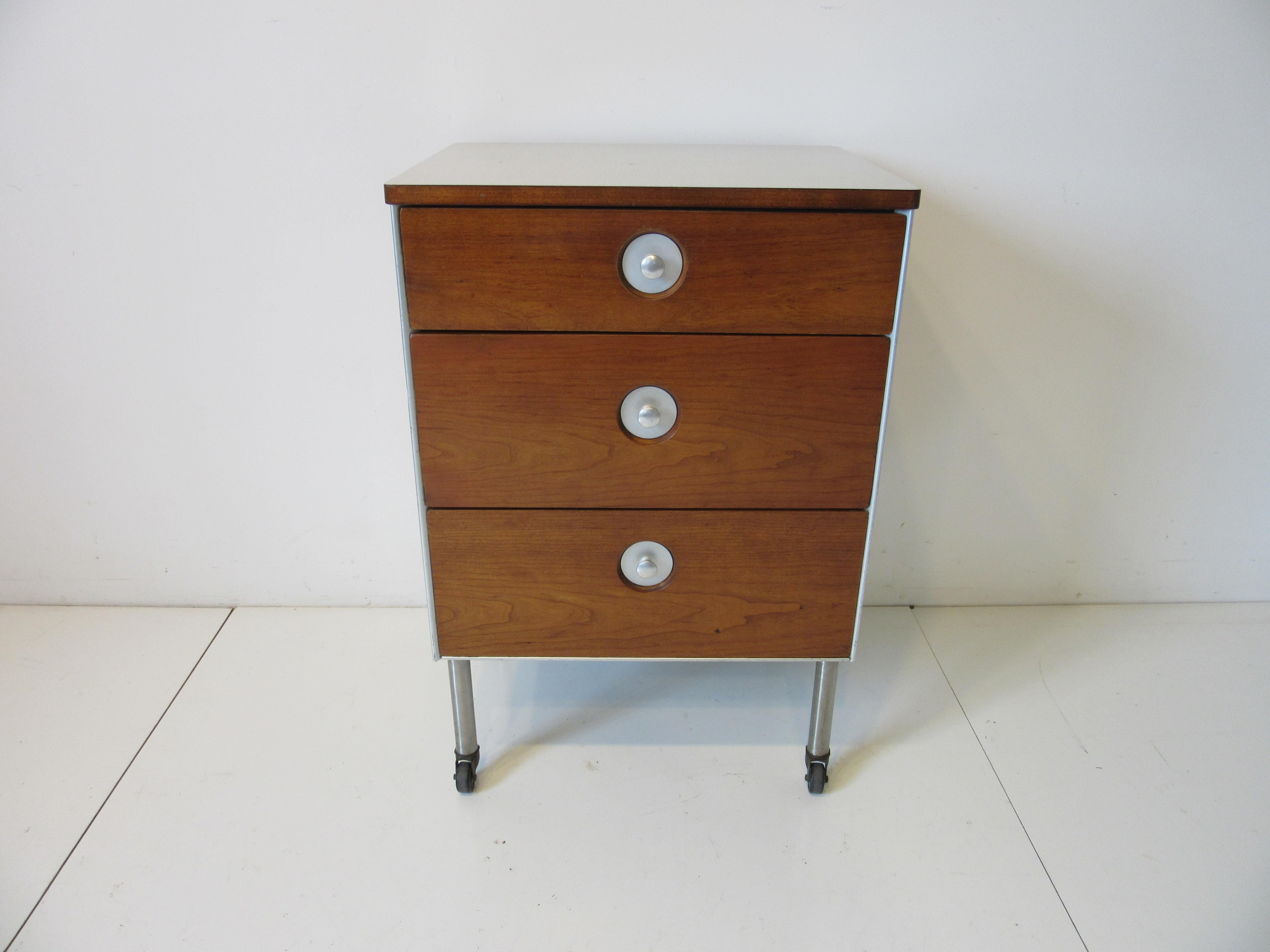 A walnut faced three drawer chest with matching sides, aluminum pulls and construction sitting on stainless steel legs with hard rubber wheels. The top is in a light grayish white toned Formica perfect for use in a bathroom or as a nightstand with a
