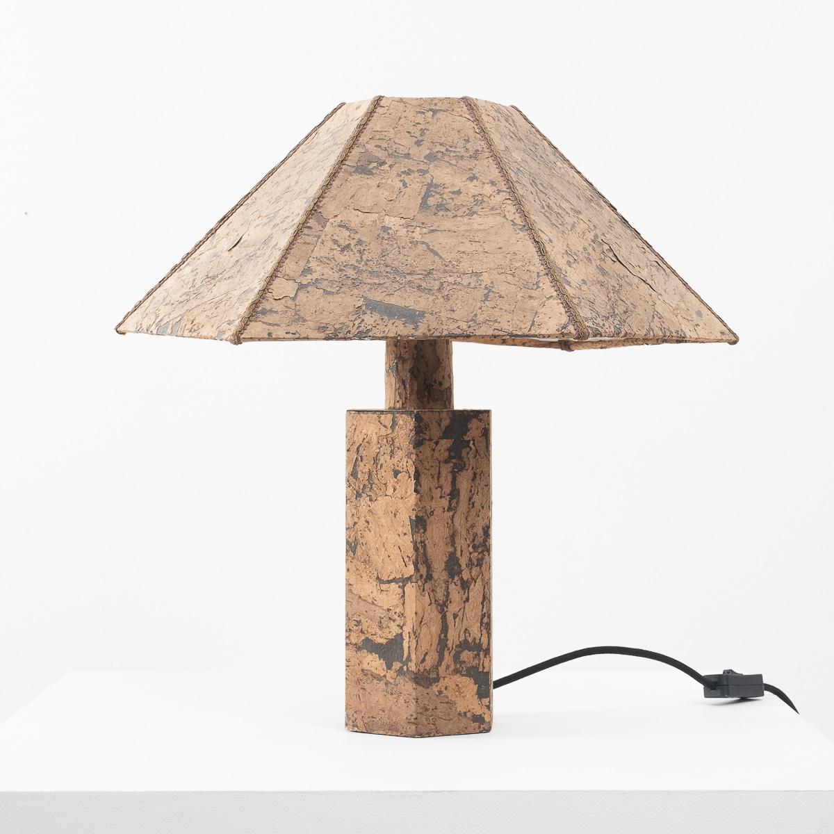 A pleasing hexagonal table lamp made from cork veneer. Redolent of a design by Wilhelm Zanoth and Ingo Maurer from 1974, which was larger with mitred edges. This one is more petite with upholstery braid masking its joins. 

It is in very good