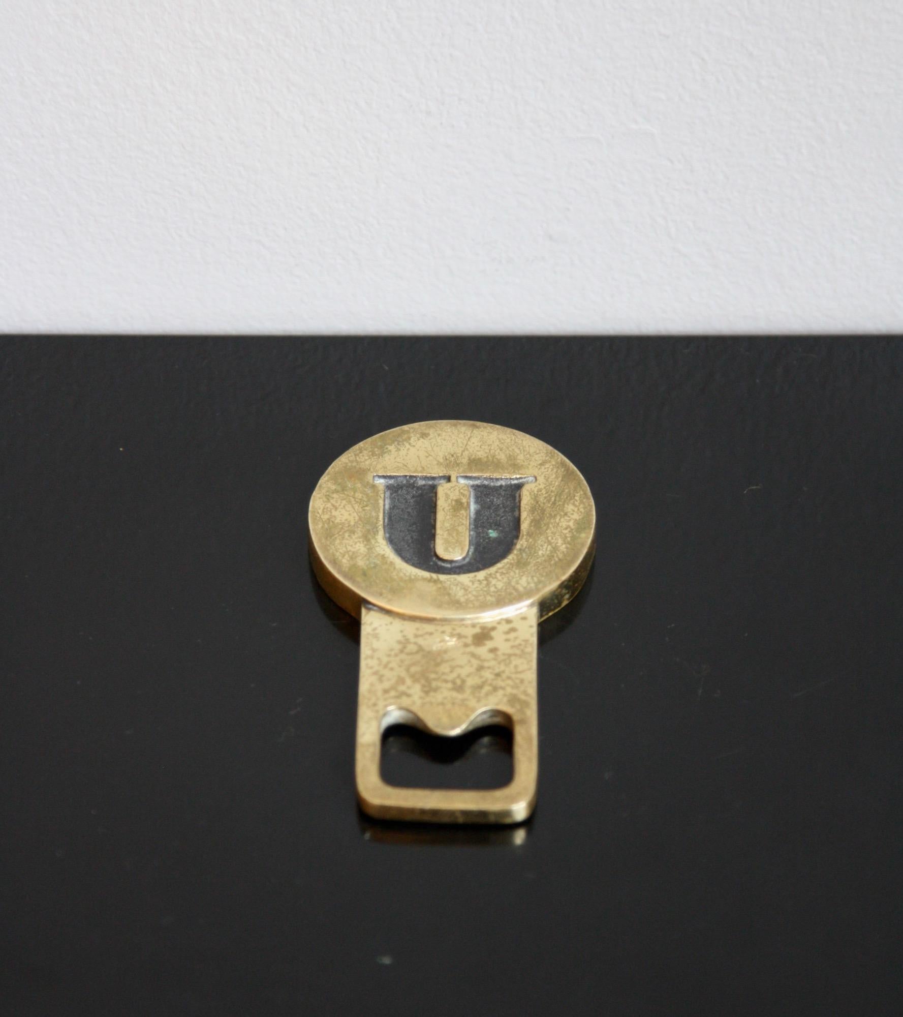 A cast brass bottle opener featuring the letter U made by Carl Auböck II, Vienna circa 1955.

The piece is formed of two geometric shapes, a circle -inside which the black patinated letter U sits- and a rectangle which houses the bottle opener's