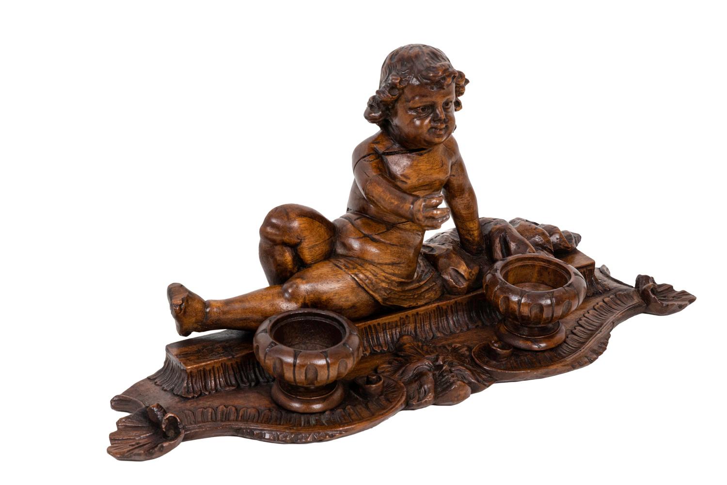 Inkwell in wood figuring a slightly lied down child only dressed with a simple cloth around his hips. He stands on a small piedestal adorned with water leaves. Two circular fluted inkwells in front of him. Scalloped base decorated with c-crests and