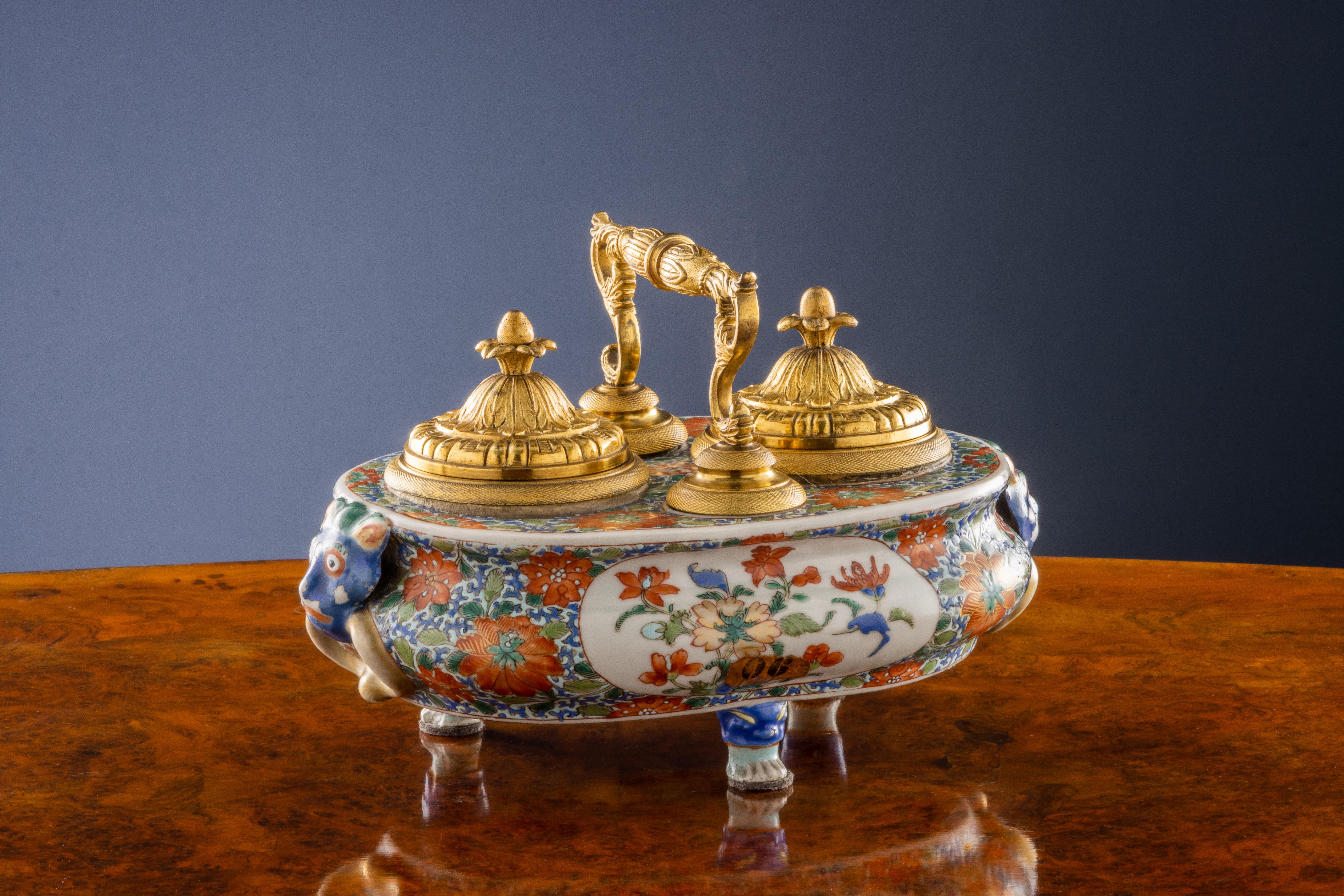 Porcelain inkwell, with a shaped body entirely enamelled with chinoiserie-style motifs: there are large red flowers on a decorated blue background, on the sides two reserves in which there is a floral composition; the inkwell rests on three feet
