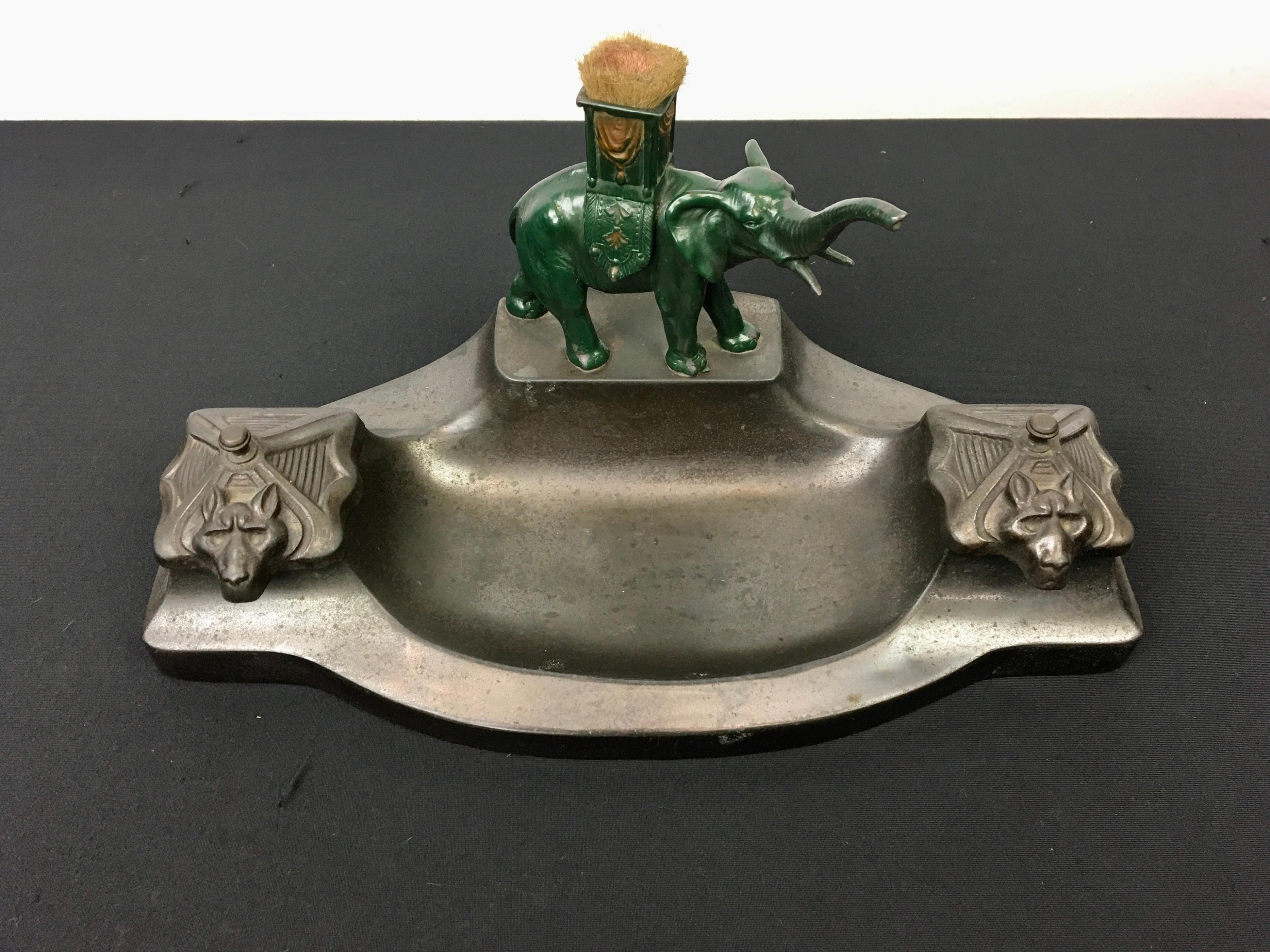 Inkwell with elephant. 
An Art Deco Inkwell with an elephant figurine on top. 
A double inkwell - double inkstand with two glass inkwells or ink containers with  reliëf lids with a kind of wolf on. In between, it's like a tray where you can put more