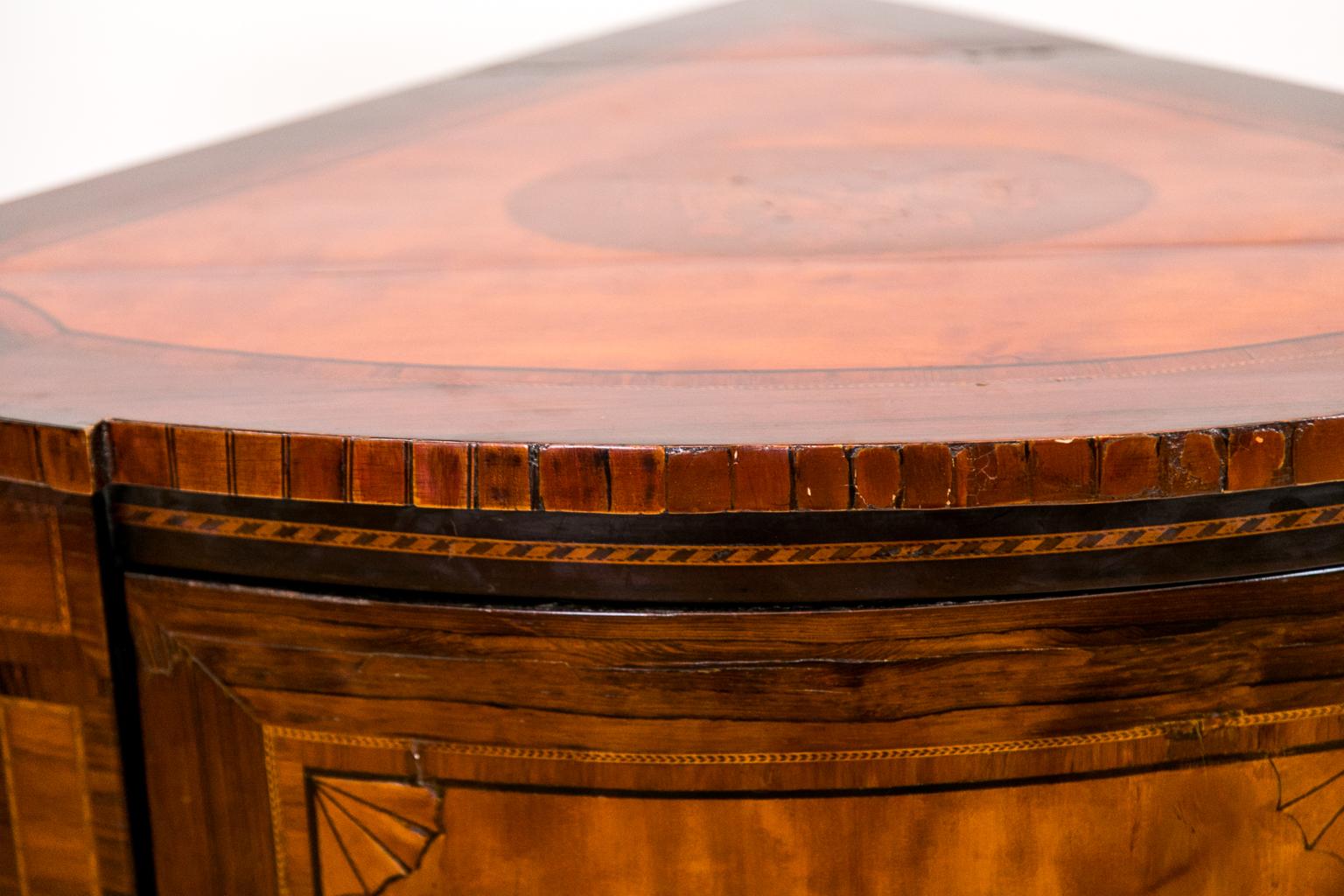 Le dessus de ce meuble d'angle à façade en arc marqueté est bordé de bois de rose avec un panneau central en bois de satin en forme d'aile d'abeille qui est incrusté d'instruments de musique. Le panneau en bois de satin est bordé d'ébène et de