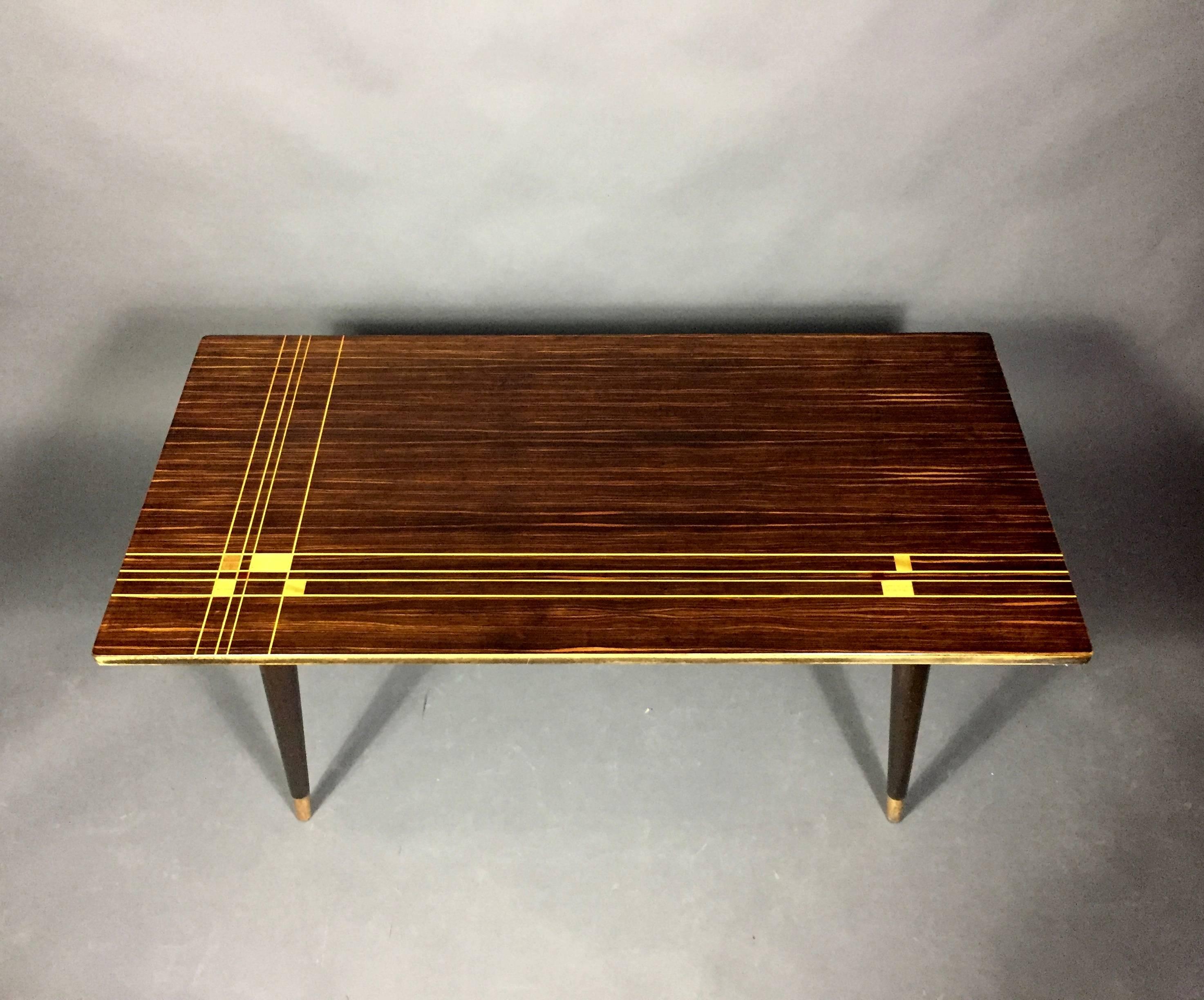 Created in the mid-1950s with a wonderful contrast top of Macassar ebony with inlaid wood in various stains in a geometric pattern. Angled legs terminate in original brass caps. Designed as 