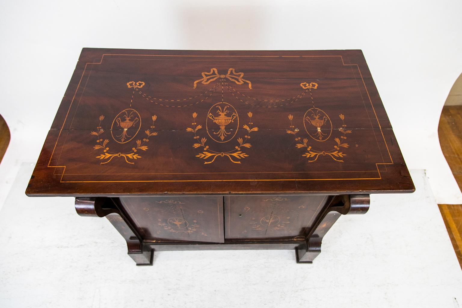 The top of this mahogany inlaid Regency console cabinet is inlaid with interlaced boxwood inlay that frames an inlaid center panel with three classical urns suspended by bow tied ribbons and stylized cords of ebony and boxwood. The three classical