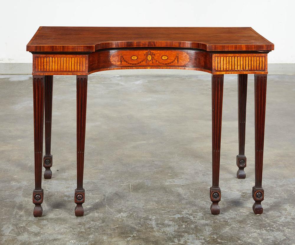 Pequeña mesa de servicio de caoba con incrustaciones, atribuida a Mayhew & Ince, circa 1775, que tiene una  La parte cóncava, con marquetería floral, descansa sobre seis patas ahusadas y acanaladas con un interesante bloqueo centrado por rosetas de