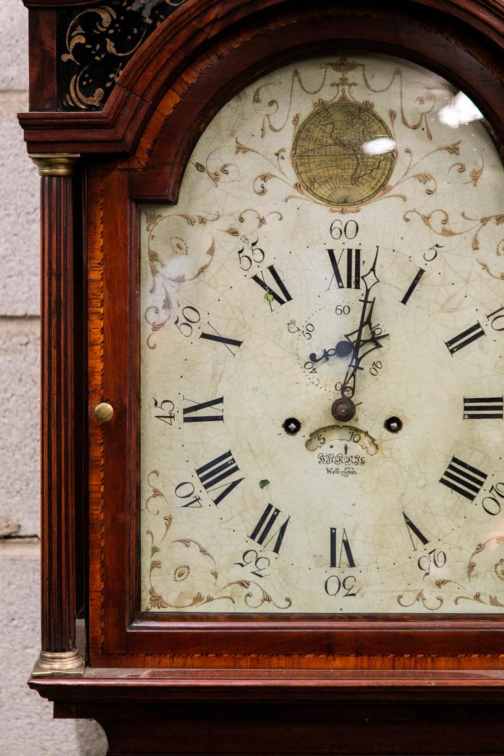 This grandfather clock has a broken arch top that has a frieze with black iglomese panels painted with floral arabesques. The right and left panels have one crack in the middle but are solid. The maker of the clock movement worked in the town of