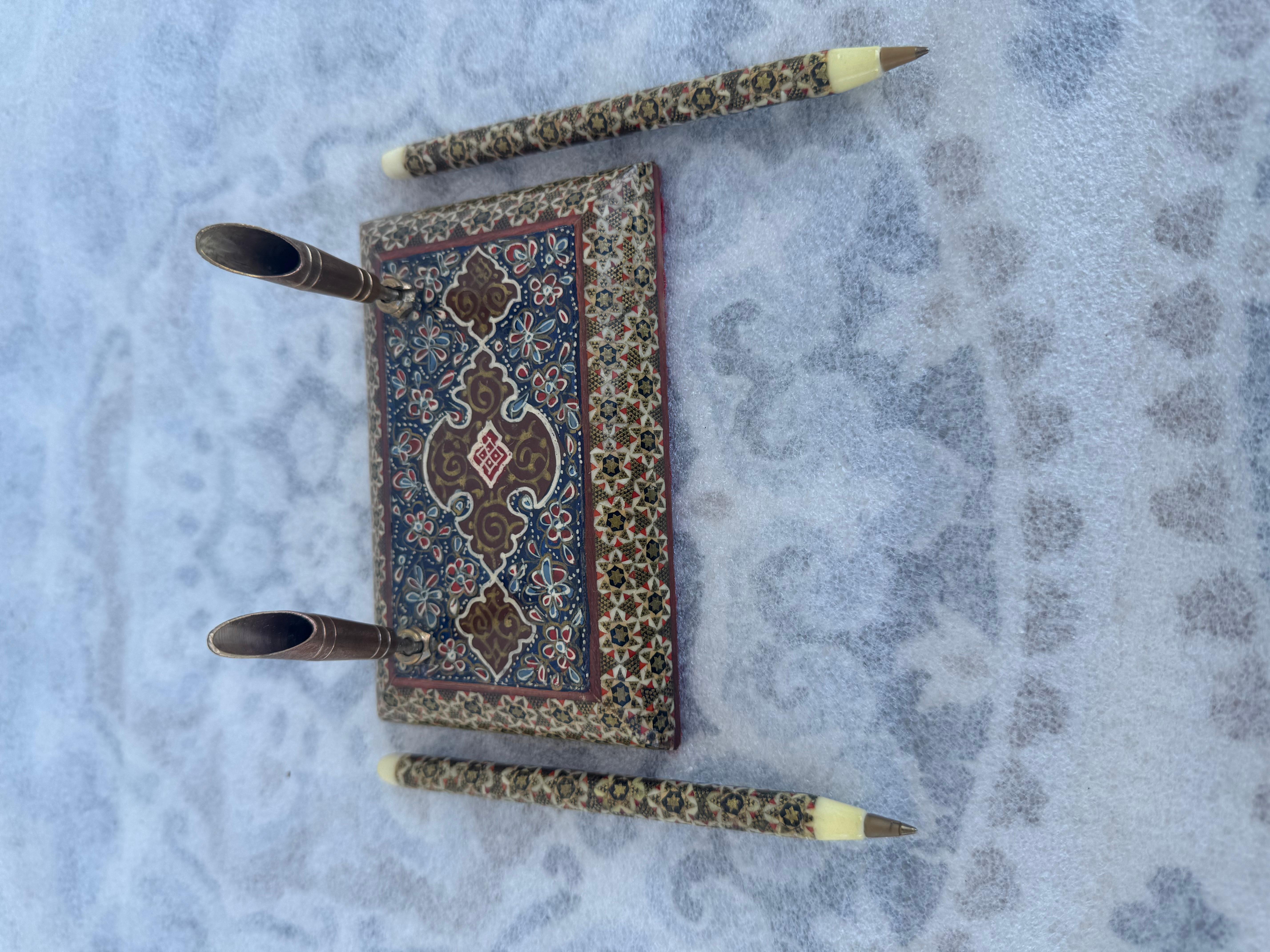 El accesorio de escritorio con incrustaciones de madera muestra la artesanía tradicional persa, con una base rectangular decorada con motivos florales y geométricos pintados a mano en tonos rojos, azules, dorados y marfil. La base está flanqueada