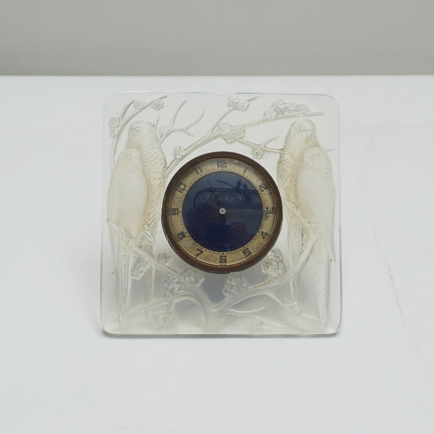Inséparables, an Art Deco glass clock. Moulded in relief with two pairs of opalescent blue lovebirds seated on prunus blossom. Dark blue cock face. Eight day Swiss movement.

Photographed on contrasting backgrounds to accurately represent