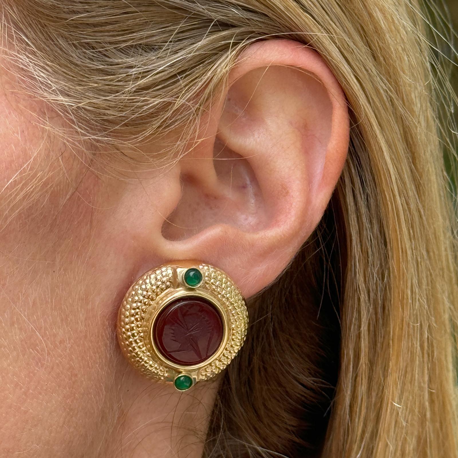 Beautiful carnelian intaglio & emerald earrings fashioned in textured 14 karat yellow gold. The round earrings feature intaglio carved carnelian gemstone flanked by emerald accents. The earrings measure 25 x 25mm and have lever-back posts. Weight: