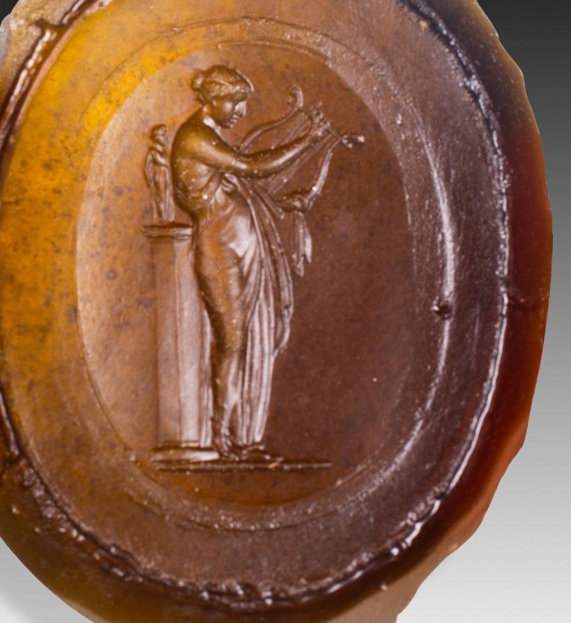  Intaglio or cameo, Muse. Glass. Possibly Italy, 19th century. 
A female figure stands, robed and carrying a harp, with a sculpture on a column behind her. This may be a Muse, so common that even Wedgewood was inspired by the base of this figure: