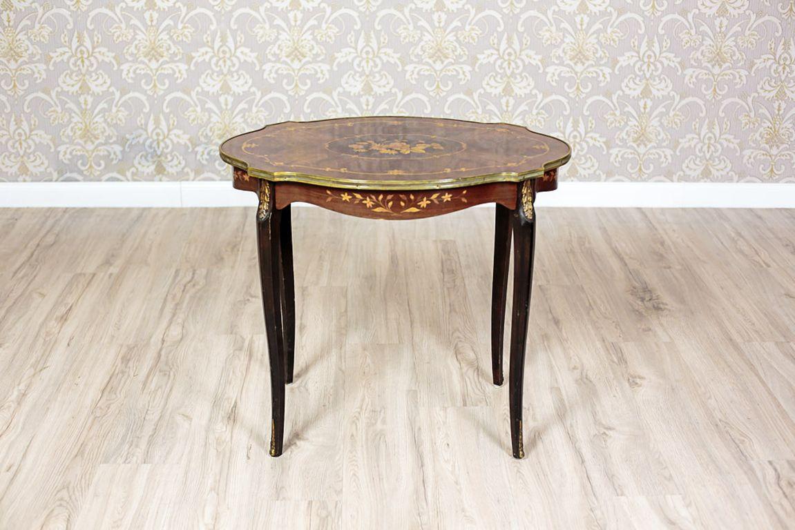 A small table from the mid-20th century.
The oval, profiled tabletop, along with the apron, are ornamented with an intarsia with a floral motif. Furthermore, the edges are finished with a brass molding.
On a side of the table, there is a drawer