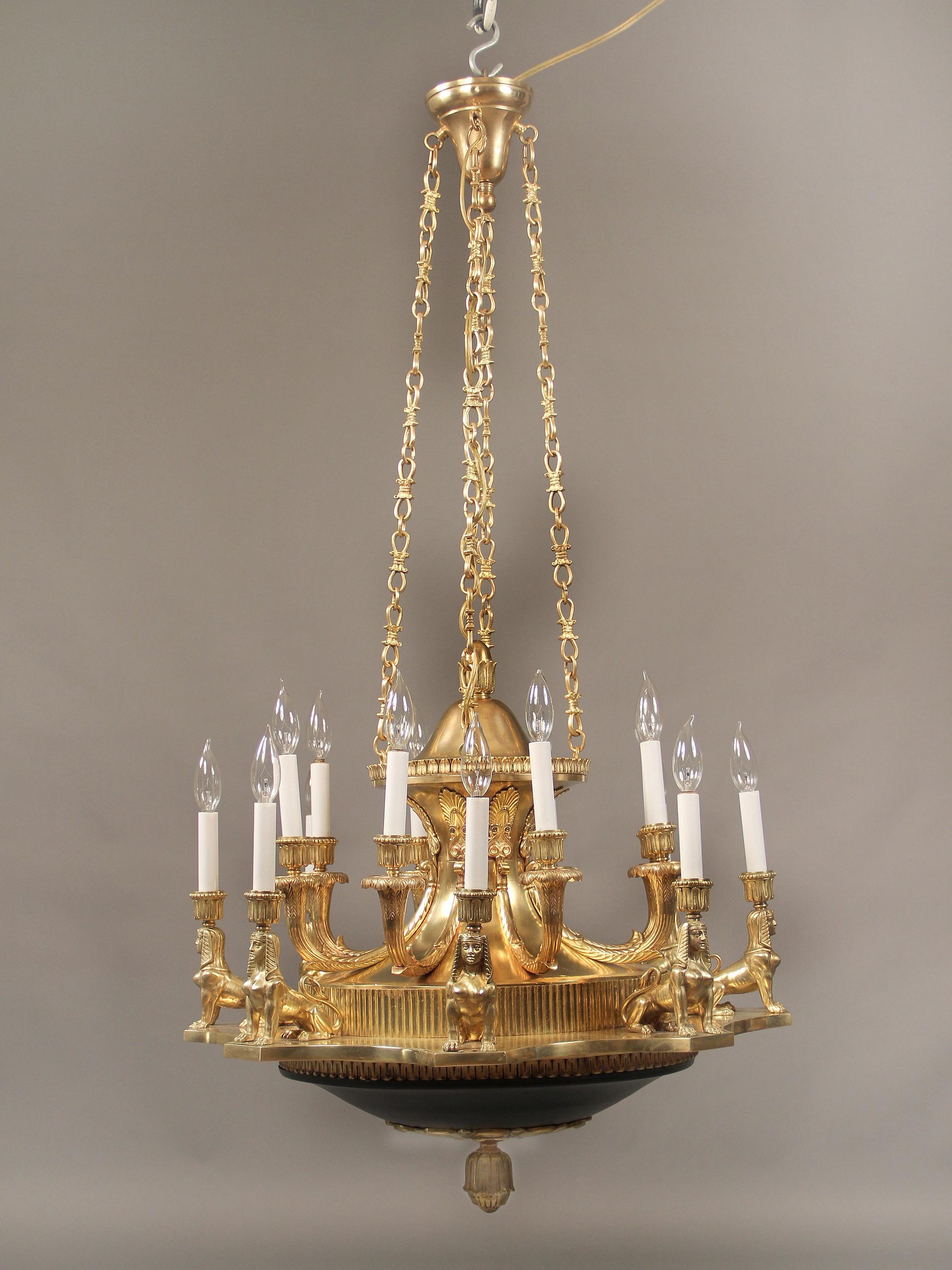 Una interesante araña de dieciséis luces de estilo imperio, de bronce dorado, de finales del siglo XIX-principios del XX

Cuatro largas cadenas unen la parte superior con el cuerpo, ocho esfinges doradas rodean e iluminan el exterior junto con