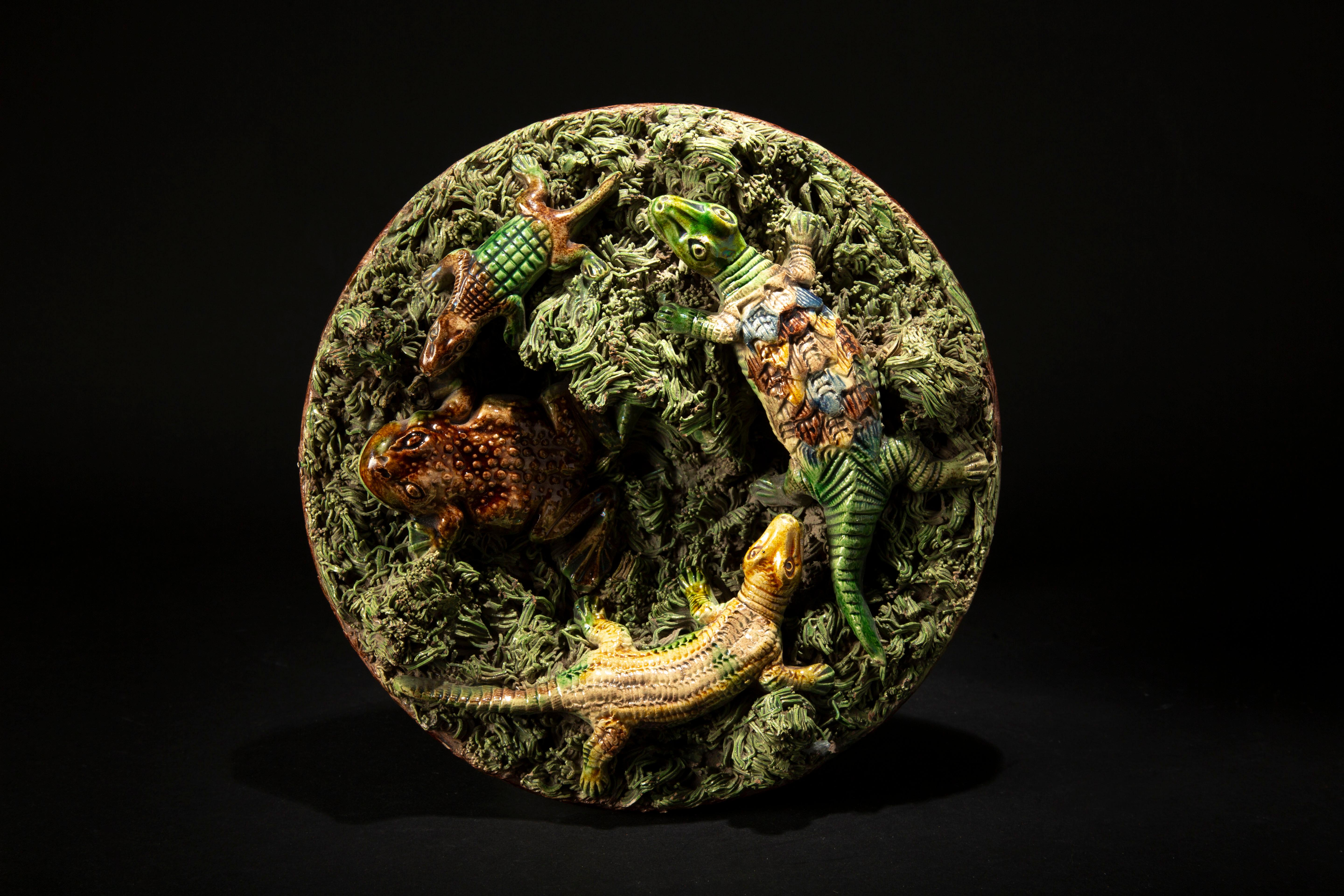 Fabricado por Caldas Da Rainha, Portugal, 1879-1885. Este plato circular de loza vidriada presenta una intrincada decoración en relieve que representa a tres lagartos y una rana en medio de un lecho de líquenes. Creado por José Alvares da Cunha