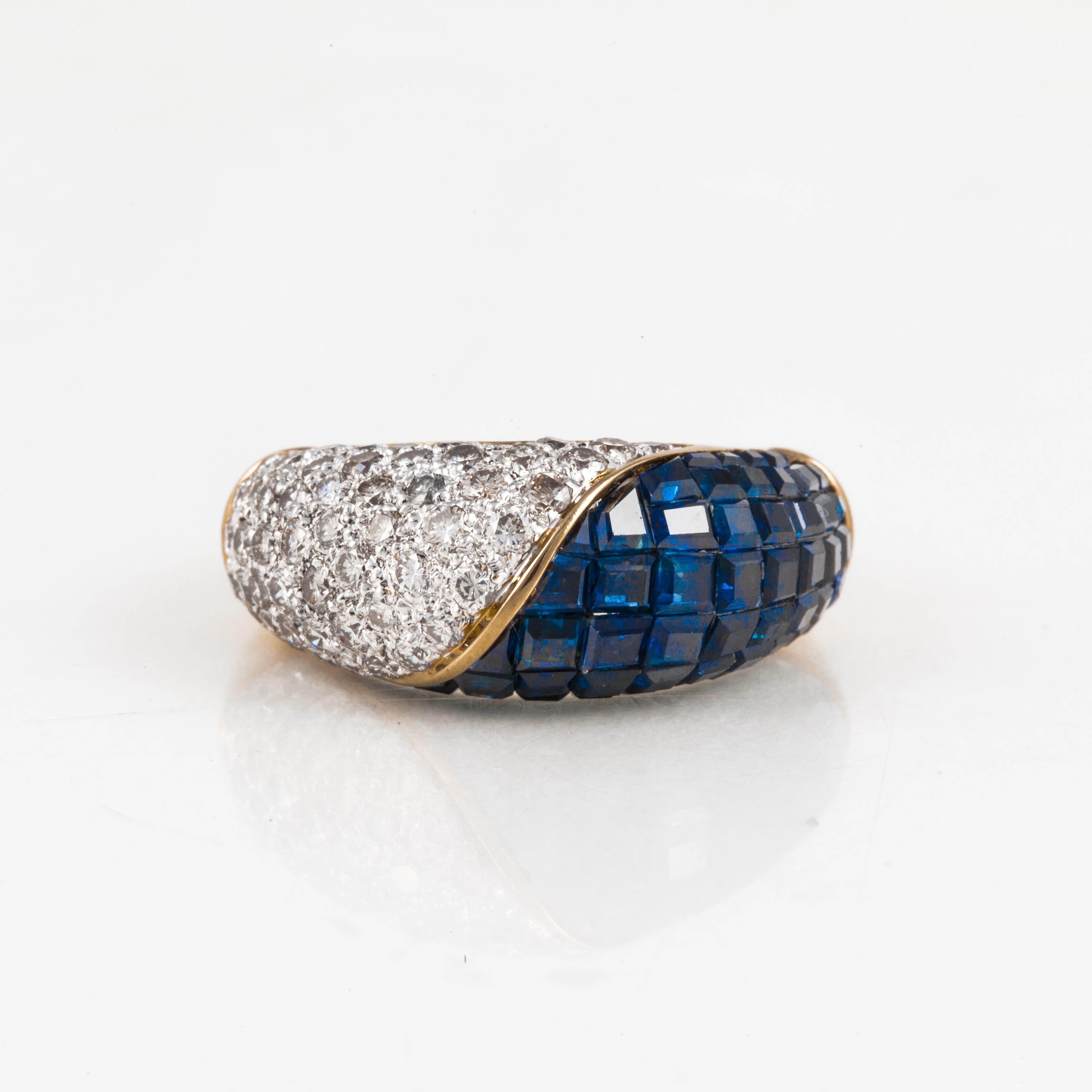 18K yellow gold ring with diamonds and blue sapphires.  There are 52 round diamonds that total 1.30 carats; H-I color and VS-SI clarity.  In addition, there are 54 square-cut invisible-set blue sapphires. It measures 7/8 inches by 3/8 inches and
