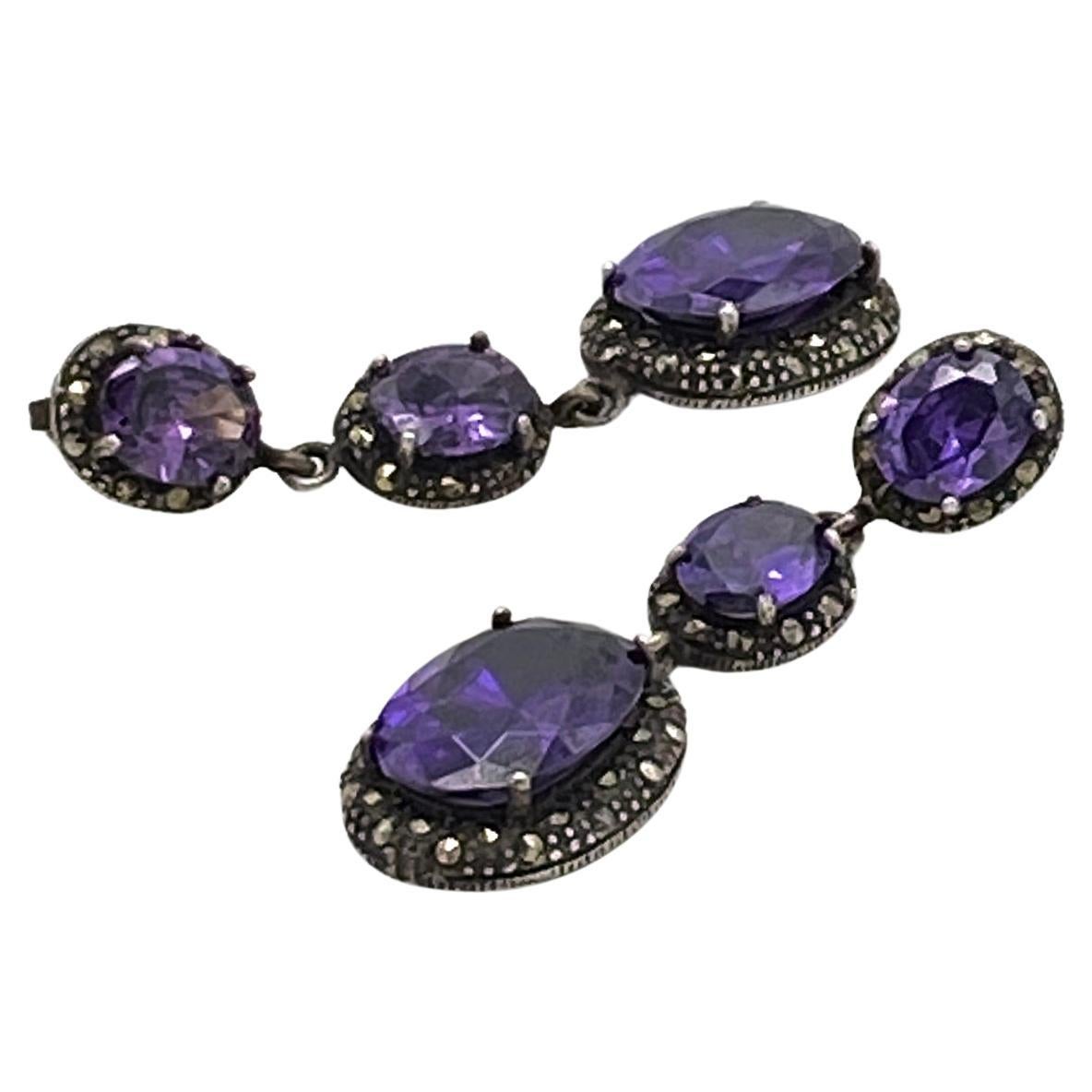 This is a pair of iolite crystal and sterling dangle earrings. There are three linked various sized oval cut transparent iolite crystal stones set in marcasite frames. These deep purple stones with dark acidified sterling frames broadcast mysterious