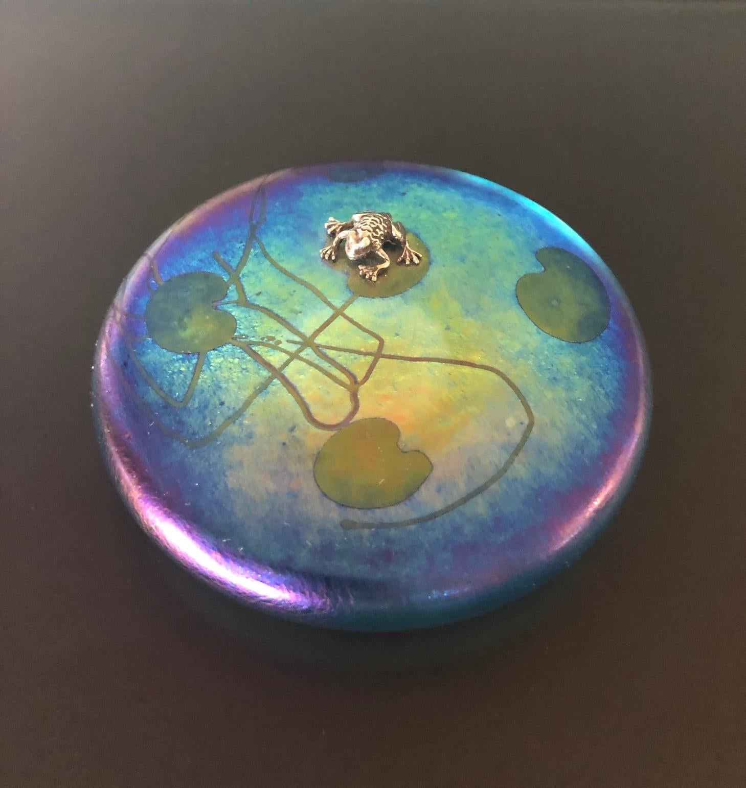 Gorgeous iridescent art glass paperweight by John Ditchfield / Glasform, circa 2000s. The paperweight of compressed circular form decorated with an applied silver frog over a purple iridescent ground with stylized lily pad leaves and whiplash lines.