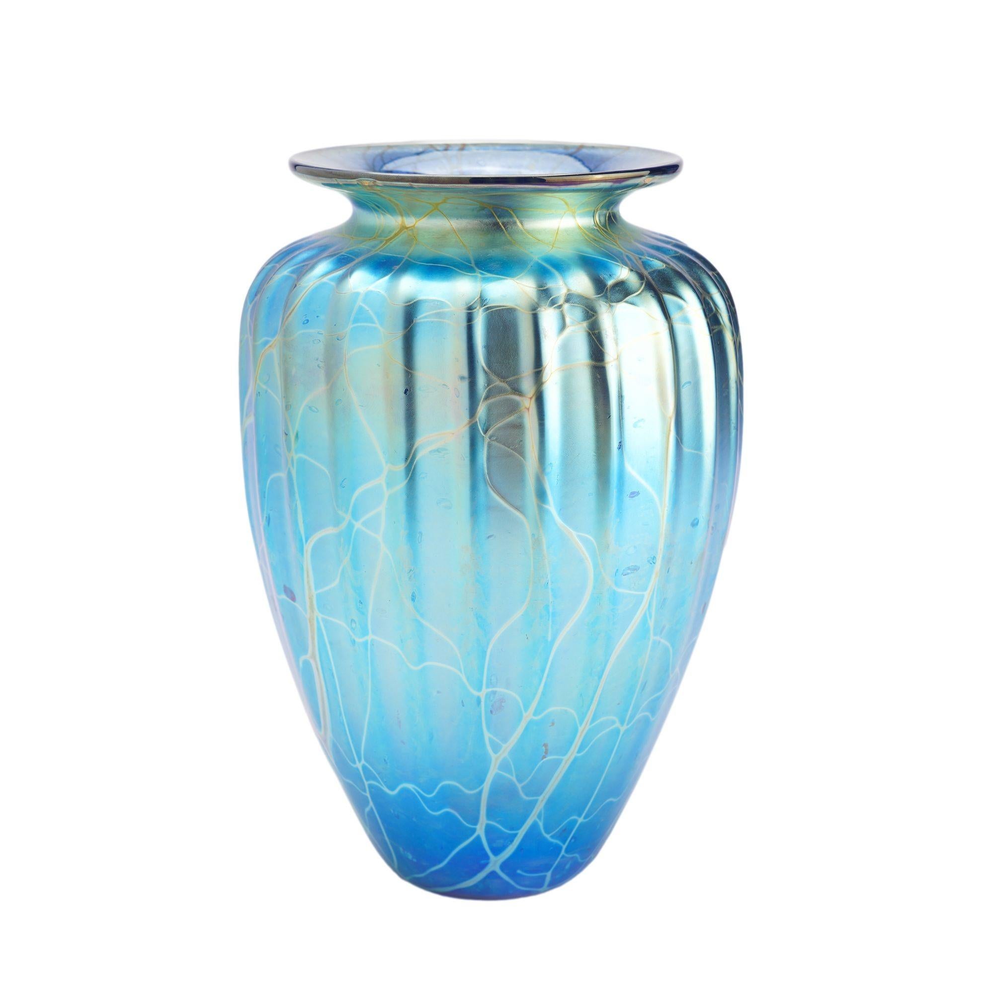 Iridescent blown glass vase with a tapered and fluted body, and waisted, flaring rim. The iridescence fades from a deep metallic blue-green at the top to a rich blue-purple at the bottom of the vase. Undertones of green, brown, and purple