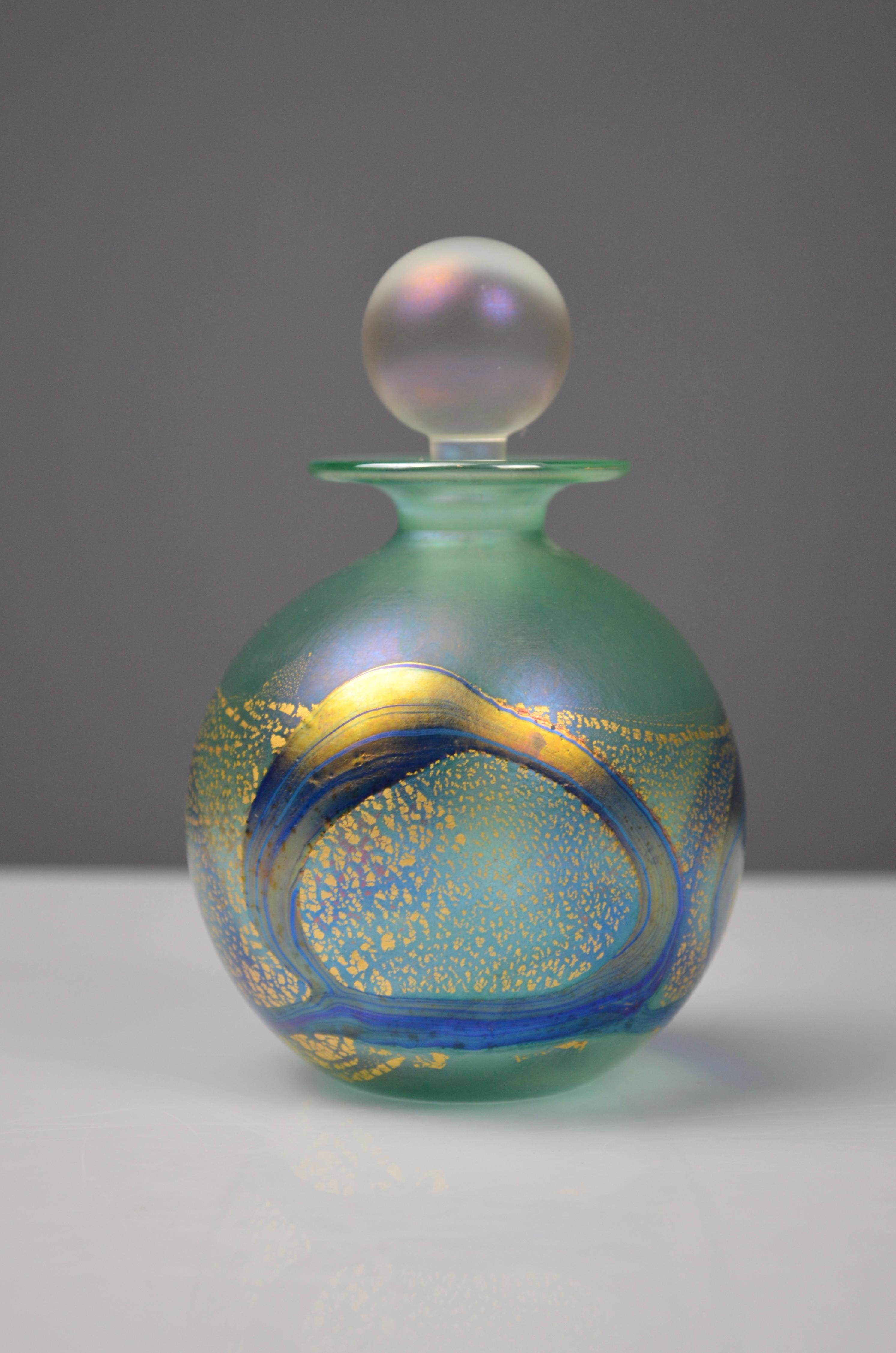 Elegante frasco de perfume de vidrio soplado de Isle of Wight Studio Glass.
El cuerpo esférico muestra una impresionante decoración verde y azul, enriquecida con remolinos iridiscentes y delicados reflejos dorados. Su tapón esférico esmerilado