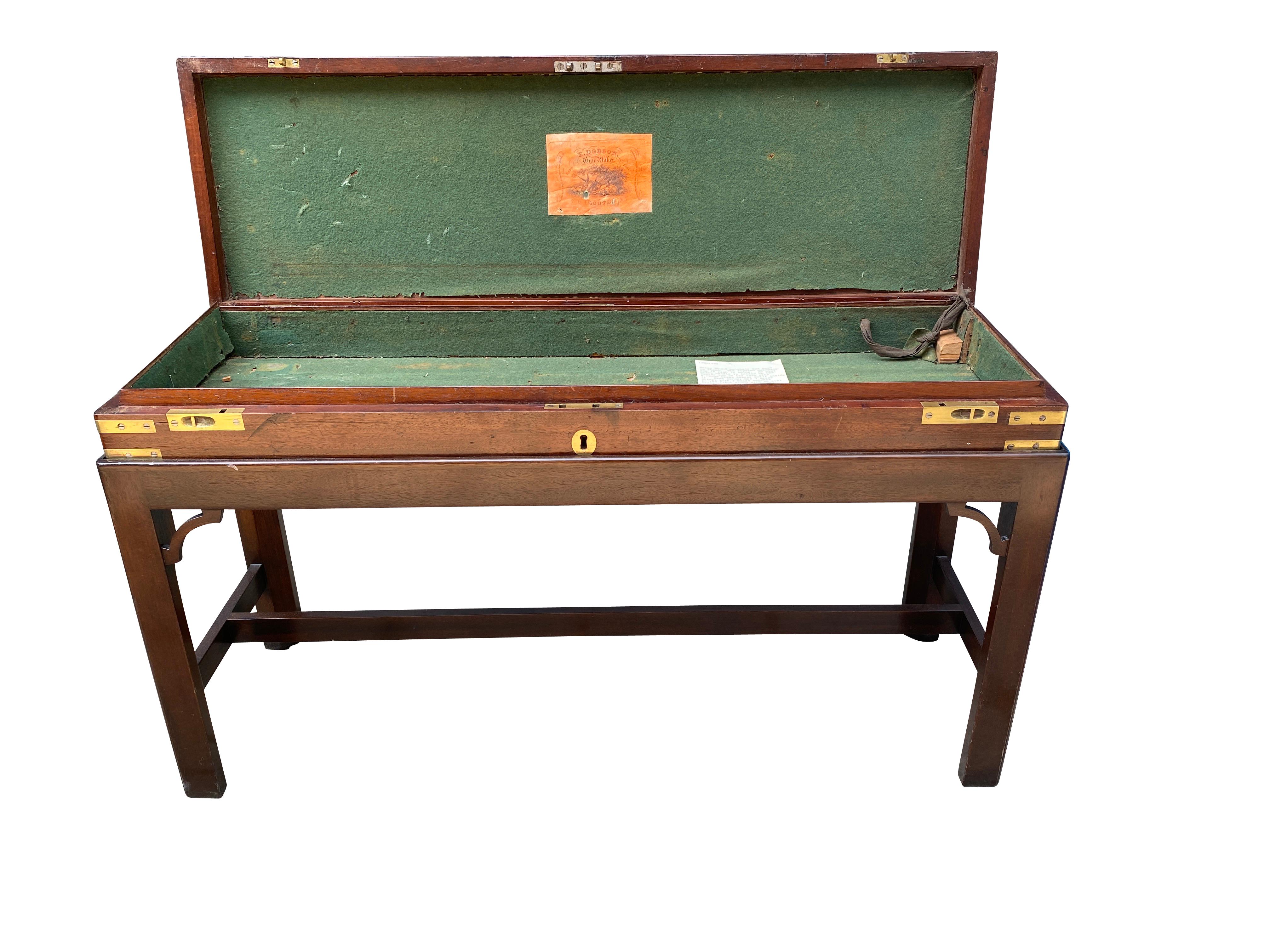 Rectangular hinged lid with central brass ring flush handle and brass corners opening to a green baize interior with a label of E.Dodson, Gunmaker, Louth. Conforming base of case set on a base with square tapered legs and H form stretcher.