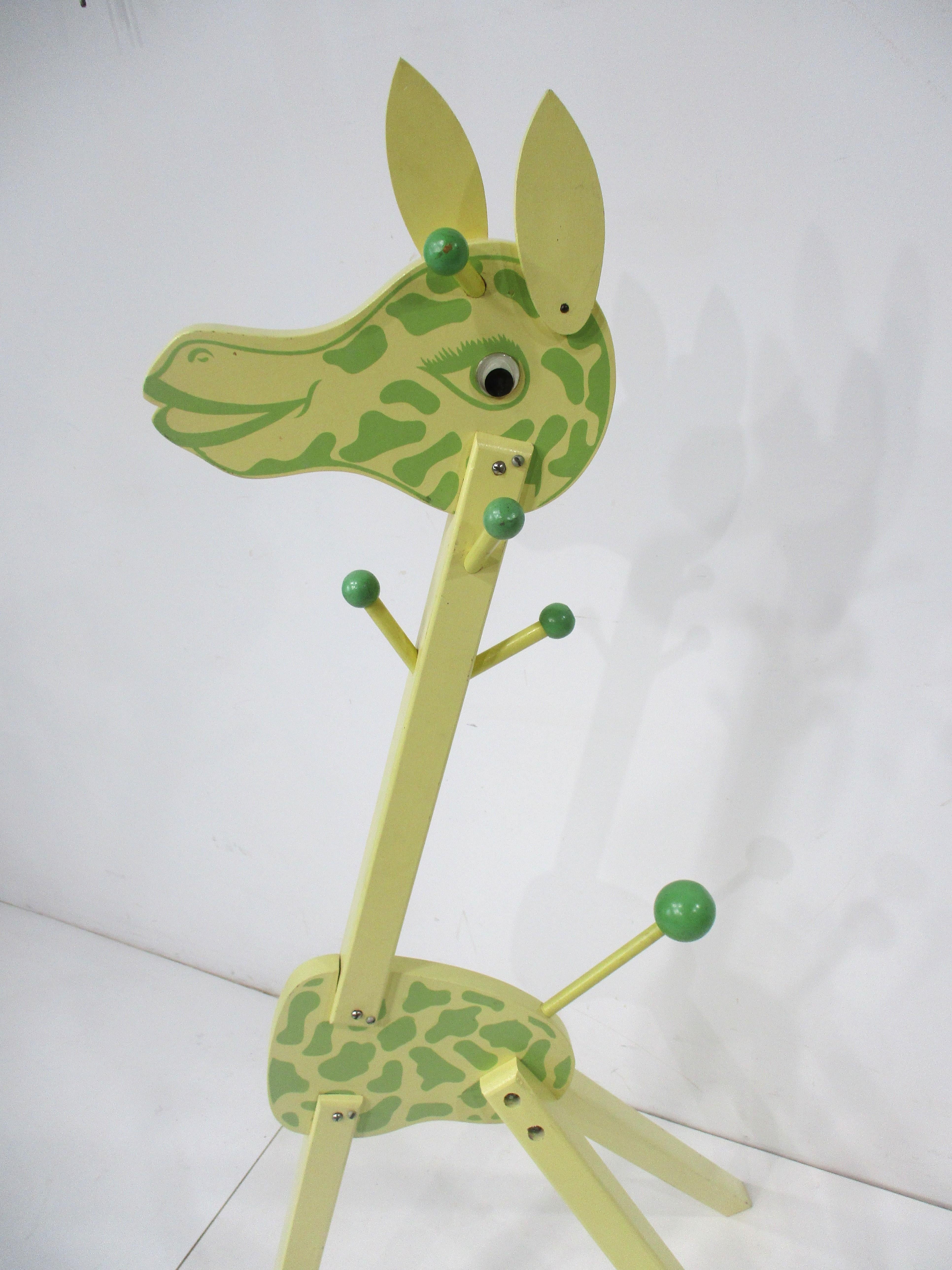 A super cute wooden giraffe childs clothing tree named Irmi in a very mellow yellow detailed with light green painted face and body spots . The body has hanger posts with contrasting colored balls and plastic eyes giving the piece great character ,
