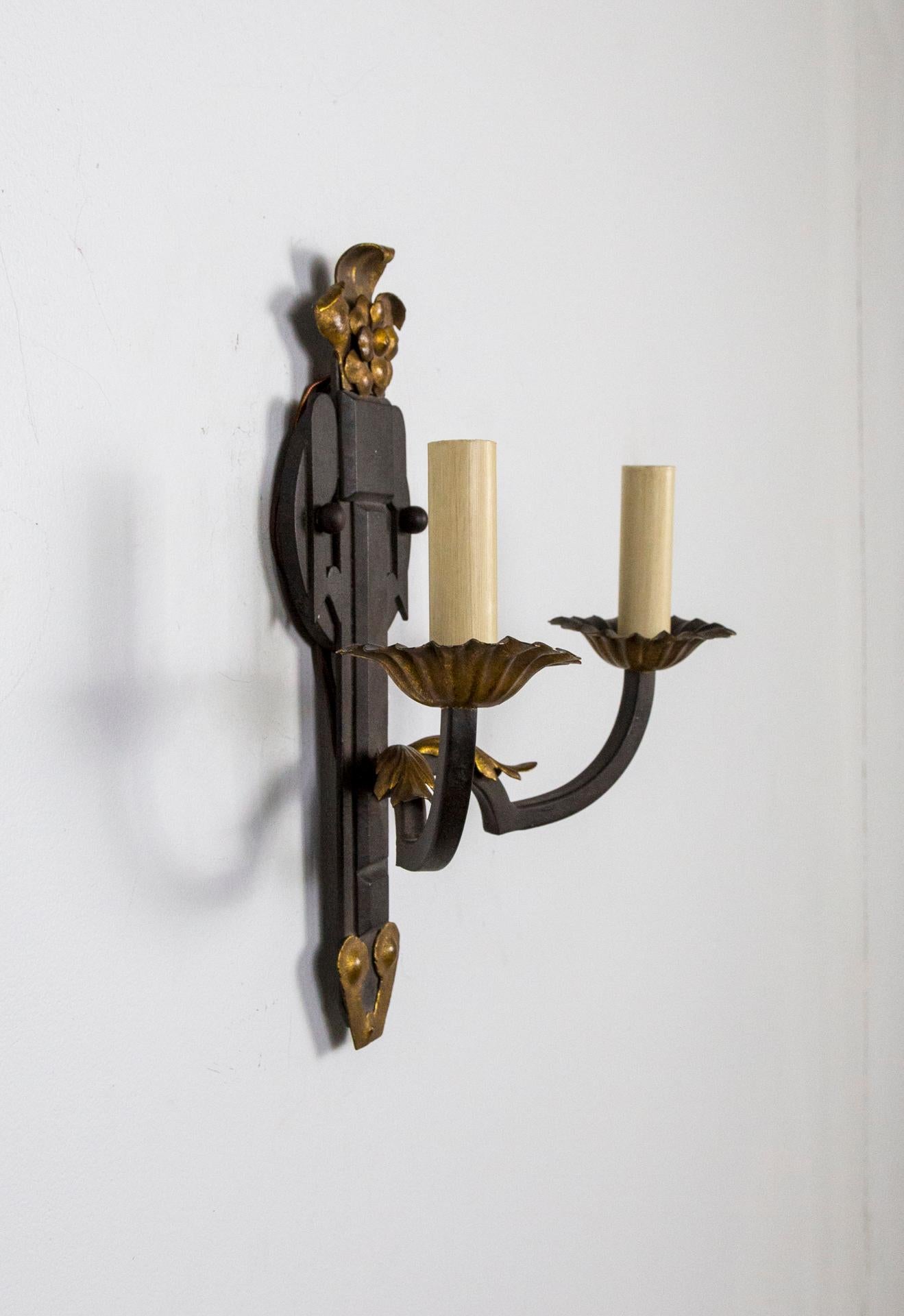 A pair of handsome, wrought-iron, 2-arm wall lights by Paul Ferrante with Deco-esque backplates adorned with a gilt flower at the tops, and gilt acanthus leaf accents.  Each arm holds a candelabra bulb.  UL listed.  14.5” height x 14.25 width x 6.25