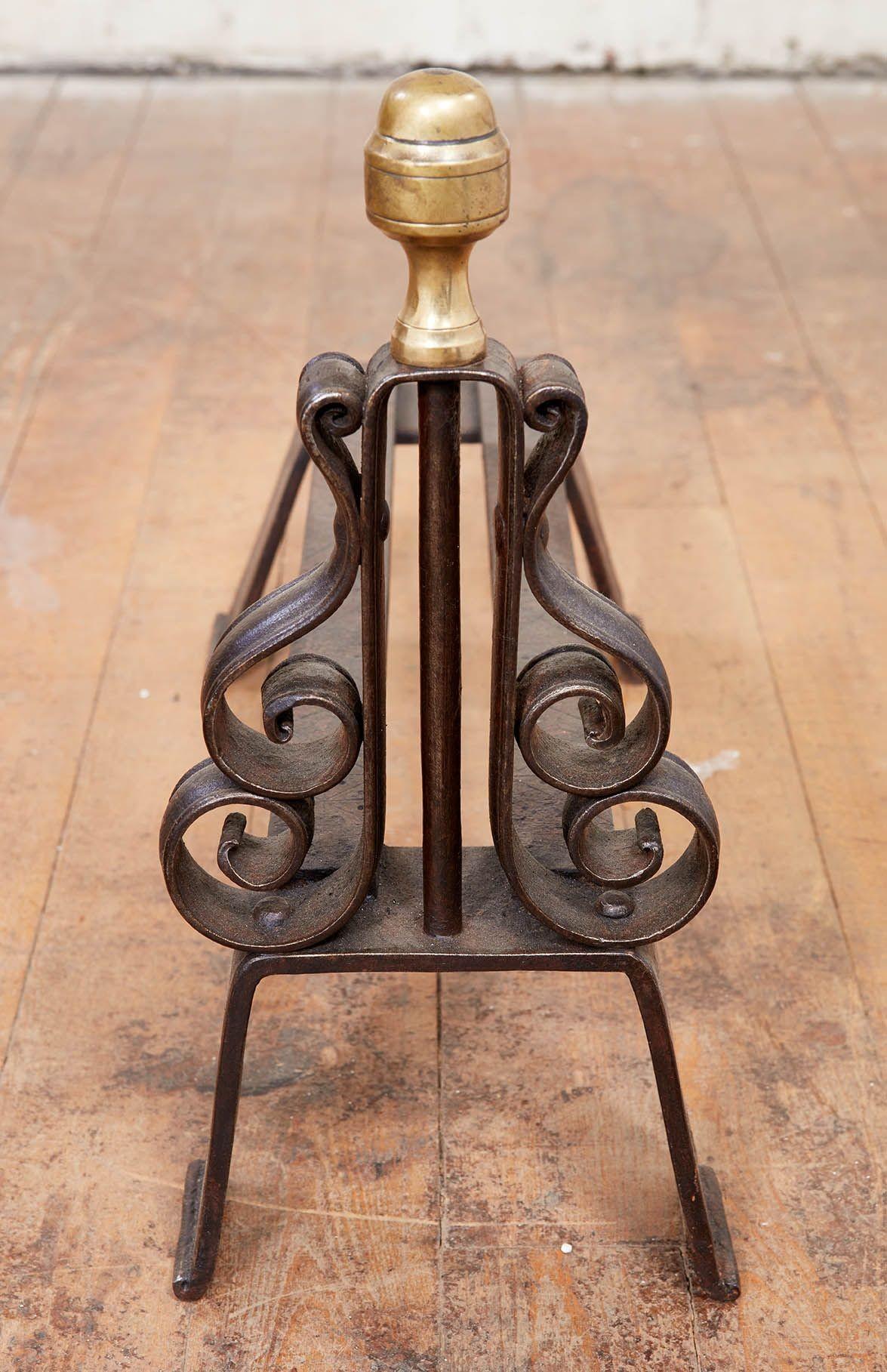 A finely-crafted pair of andirons with lyre shaped bodies in scrolled iron surmounted by a turned brass finial, the whole on two sets of inverted U strap legs joined by double rails incised with decorative line and dot markings.