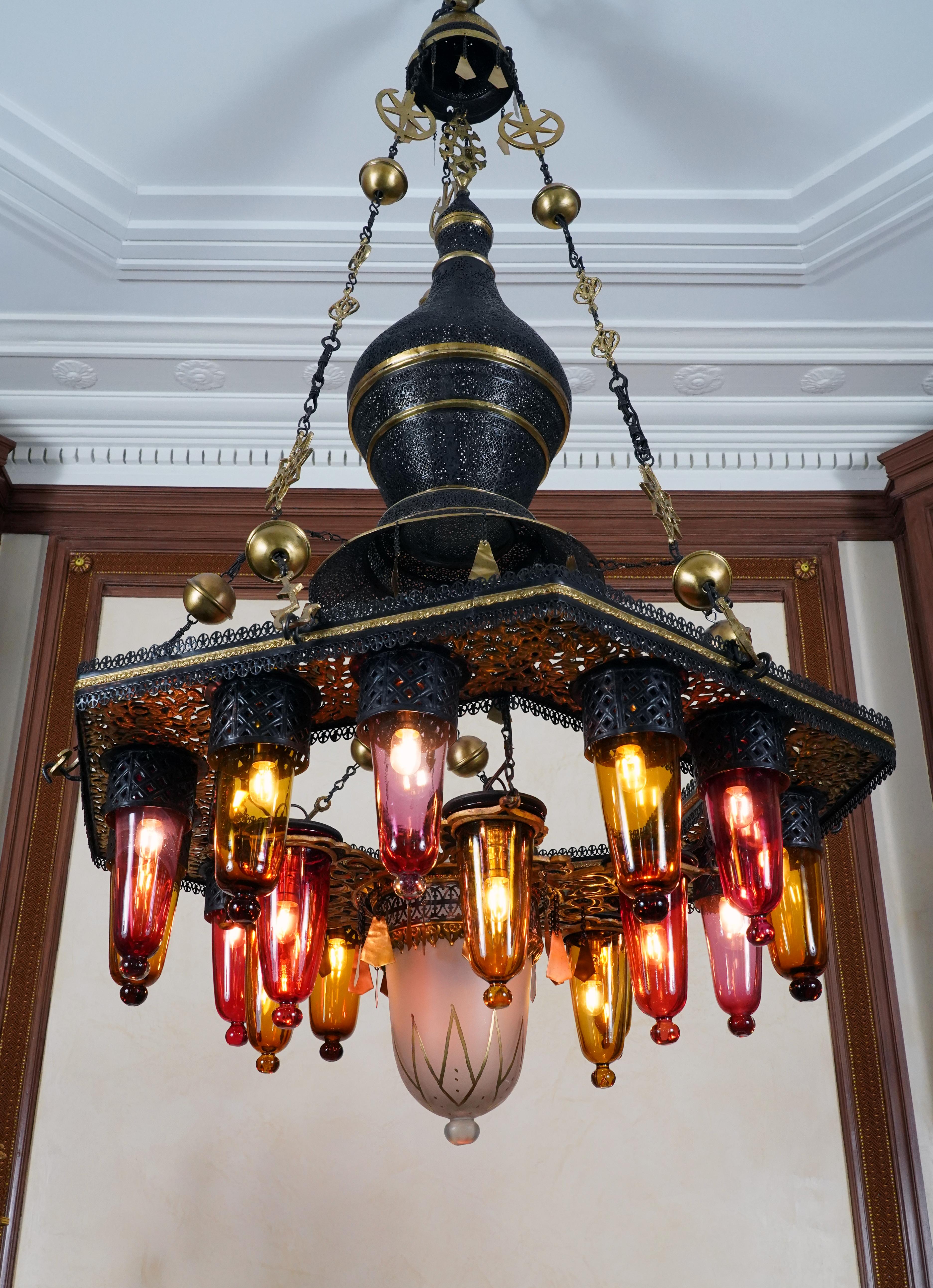 Raro lampadario marocchino in ferro traforato patinato e dorato a diciotto luci,
composto da un fusto a balaustro che termina con una corona esagonale circondata da una galleria e decorata con 12 vetri illuminanti alternati di colore ambra e rosso e