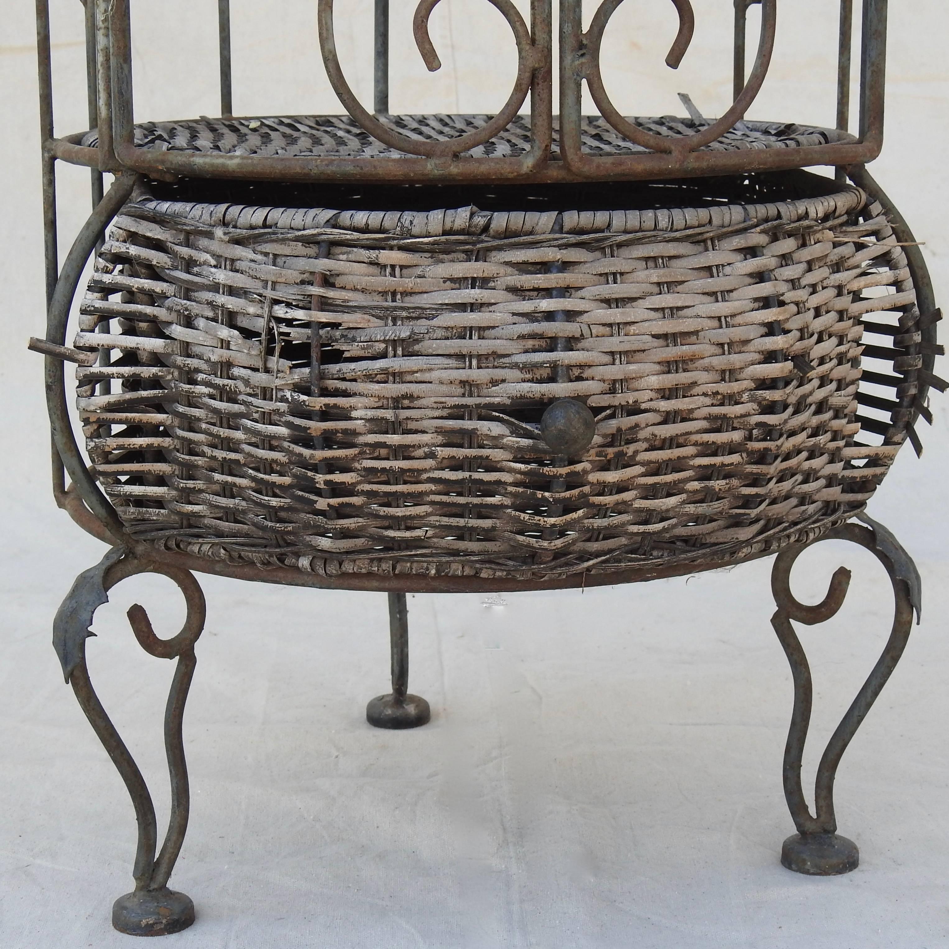 Rustic scrolled iron forms the body of this unique wine rack which will accommodate six bottles of wine behind the hinged door. There is a shelf on the top made of wicker. The bottom also has a shelf with a drawer for storage.