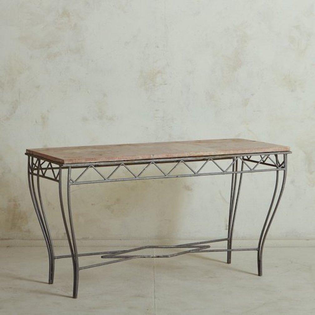 A 1960s French console table featuring an intricate wrought iron base with a gray finish and a triangular motif trim. This table has a pink marble tabletop with cream and gray veining. It stands on curved, tapered legs and has a diamond shaped