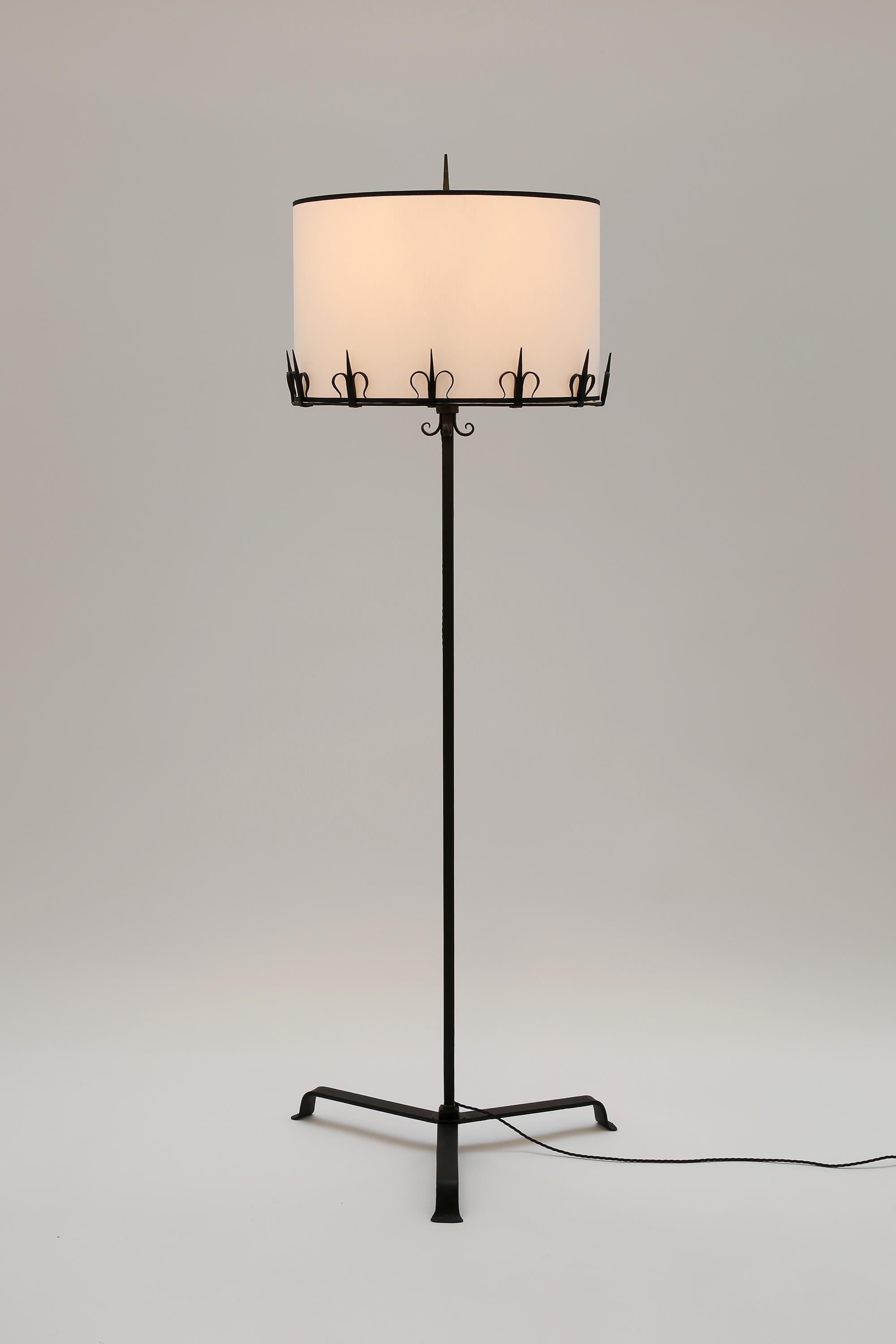 A run of five forged and enamelled iron floor lamps in the taste of Les Artisans de Marolles, with decorative fleur-de-lis detailing, three point bases and new white cotton shades with black trim. From a hotel in Andalusia, southern Spain, c. 1960.
