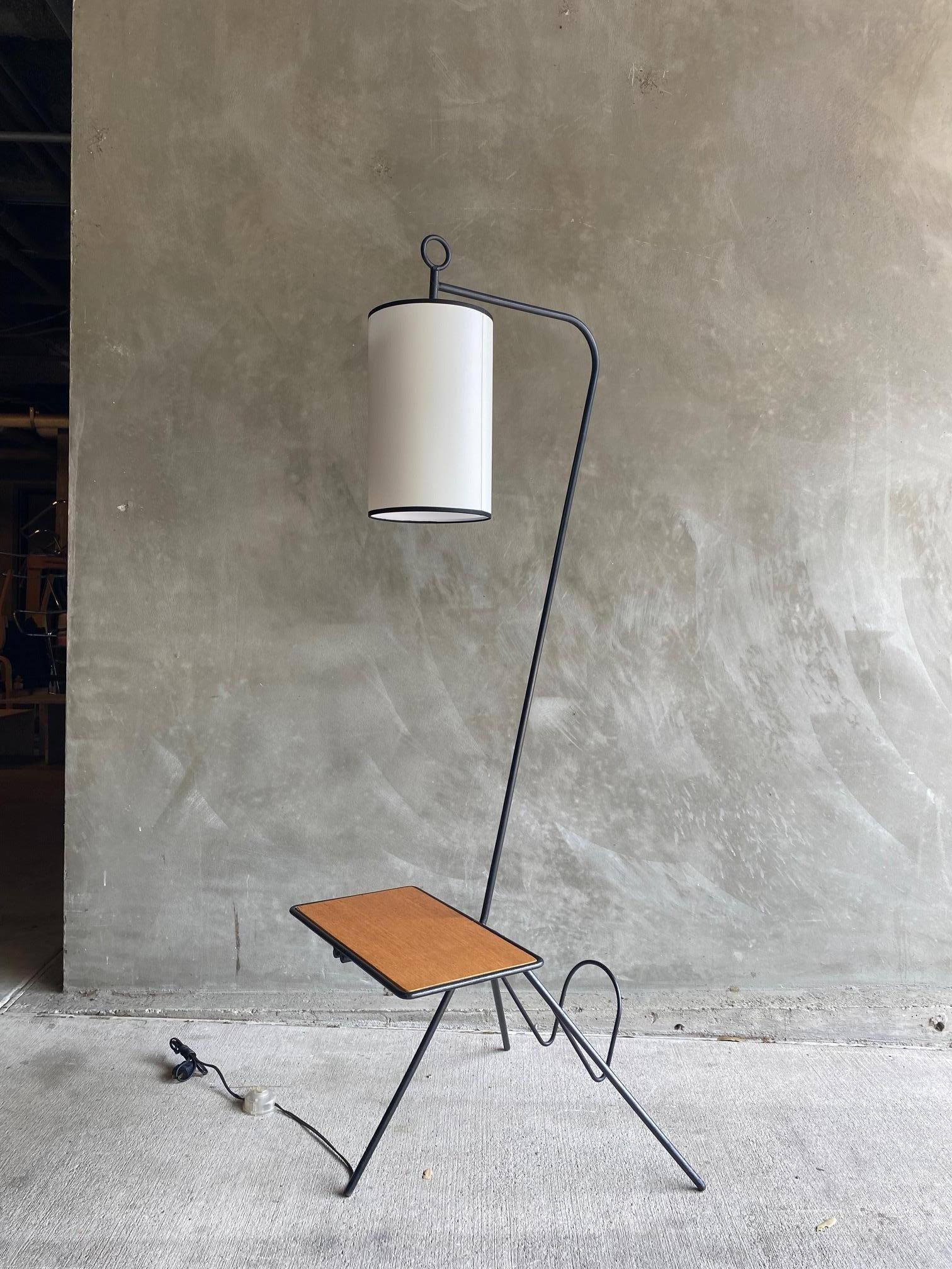 Tripod iron floor lamp with integrated table and magazine caddy. Solid iron frame with rattan detailing supports a natural wood inset table surface. Backside features a simple magazine or newspaper holder. New linen drum shade secures tightly.