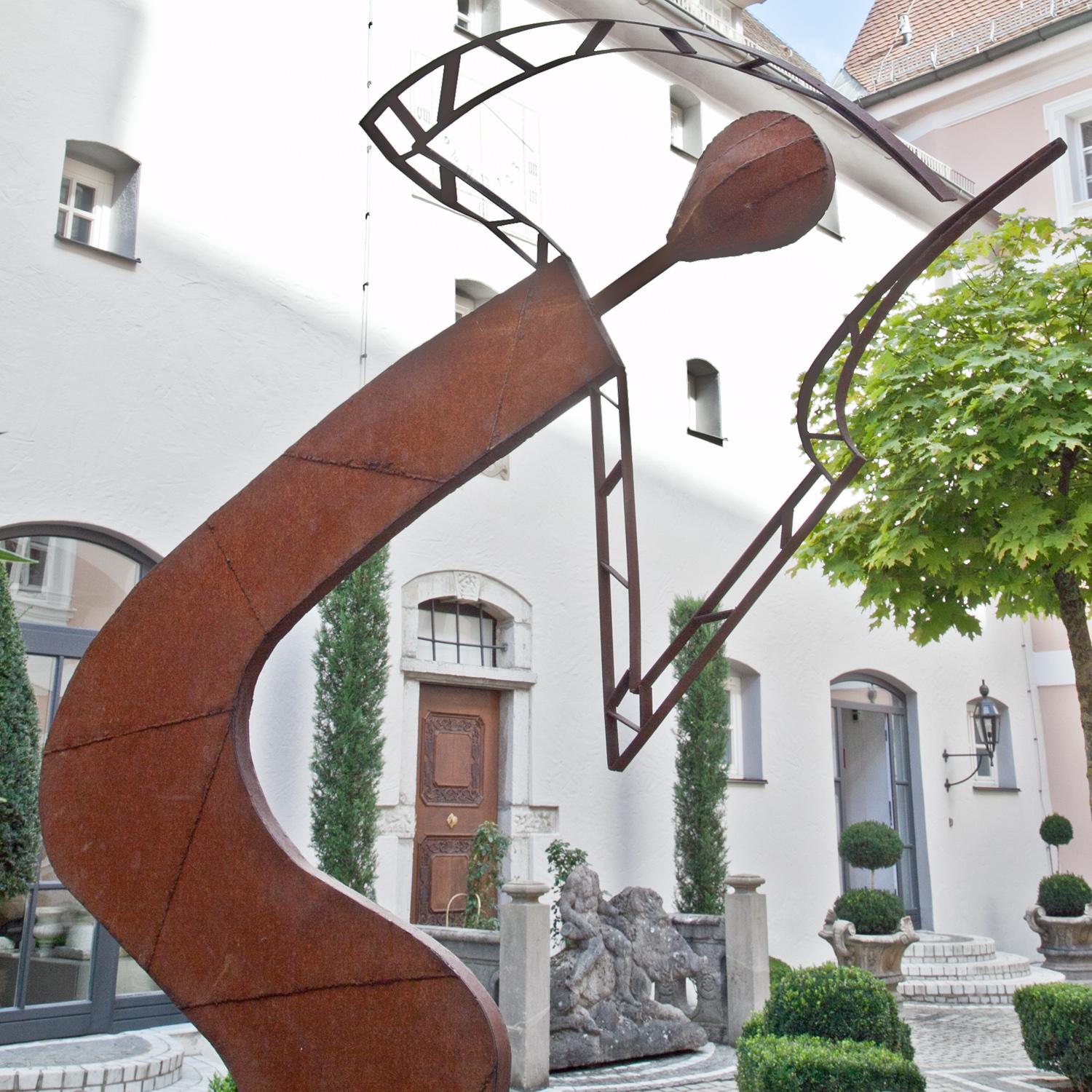 Larger than life-sized iron sculpture is mounted on a rectangular base and can be rotated. The sculpture in an abstract humanoid form has angled arms and an S-shaped body and slightly sways in the wind.