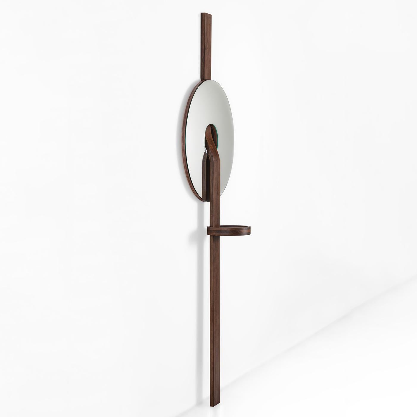 This wall mirror is a triumph of geometric volumes arranged in a sculptural composition of unmistakable contemporary inspiration. The disk-shaped mirrored glass element is pierced by the slightly curved band in solid Canaletto walnut that serves an