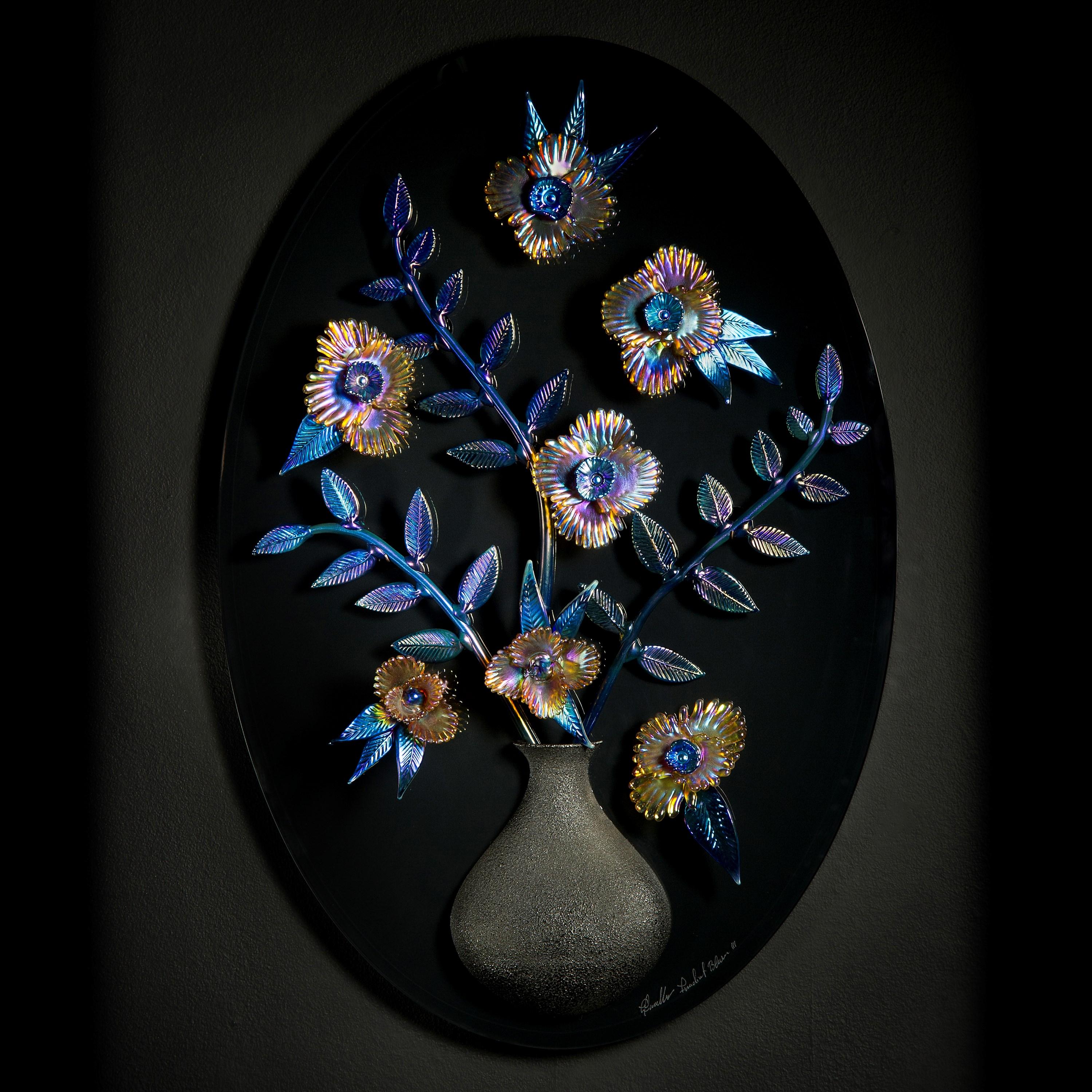 Irradiant Bloom III is an oval sculptural wall mounted artwork by the British artist and Blown Away II series winner, Elliot Walker. The artwork features hand-sculpted iridescent flowers with a bisected black vase that is bonded to the back. The